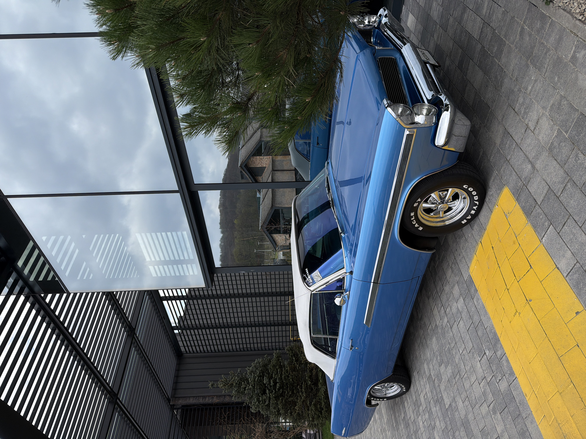 A light blue classic convertible car with a white soft top is parked on gray paver stones, partially obscuring a bright yellow painted stripe on the ground. The car features chrome bumpers, a prominent grille, and chrome wheels fitted with "GOOD YEAR EAGLE ST" tires. A partially visible Ukrainian license plate, possibly reading "U404BU", is on the front. On the rear side window, a sticker with text including "DESERT RACE" and "CHOPTOP" is visible. Behind the main car, another dark blue or purple car is partially visible.
The setting appears to be an outdoor area next to a modern building with extensive glass windows and a distinctive dark structural facade, possibly a pergola or roof. The glass reflects the overcast sky and elements of the surroundings. Lush green pine trees are prominent in the foreground and midground, casting shadows. In the background, beyond the building, hills or mountains are visible under a cloudy sky. The overall weather is overcast and cloudy, indicating daytime. The scene is located in Vynnyky, Ukraine.