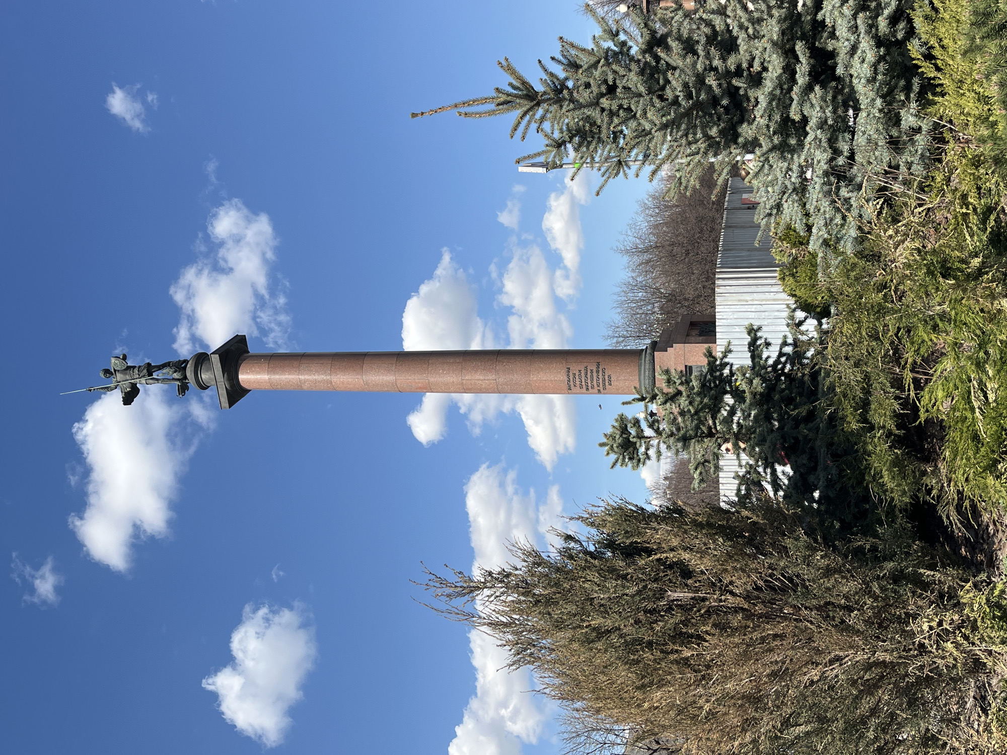 The image shows the Alexander Column in Moscow, Russia, set against a blue sky with scattered white clouds. The column, made of red granite, has a dark pedestal and a bronze statue on top. Various trees and bushes surround the base of the column. A metal fence is visible behind the greenery. The image appears to be taken on a sunny day.