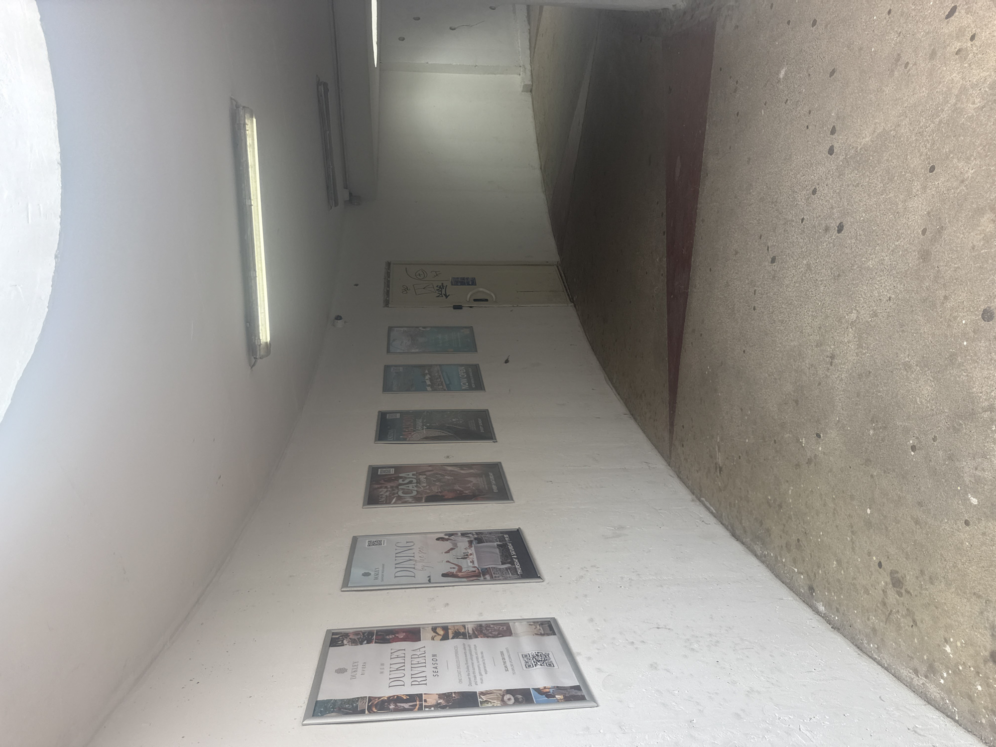 This media file depicts an indoor passageway, likely a corridor or tunnel, in Bečići, Montenegro. The scene is characterized by a stark white, curved wall on the left and a rough, textured concrete wall and floor on the right. The lighting is artificial, with fluorescent tubes visible along the ceiling, suggesting it is not daytime.
Along the white wall, a series of framed posters or advertisements are displayed vertically. These posters advertise a place called "DUKLEY RIVIERA," with specific mentions of "DINING by the sea," "CASA," and "SEASON." One poster prominently features the text "DUKLEY RIVIERA NEW." Another poster indicates "THURSDAY & SATURDAY 19:30" and "EVERY SATURDAY." A QR code is also visible on one of the posters. Further down the corridor, a closed door is visible, adorned with graffiti.
The overall impression is one of a somewhat utilitarian but potentially upscale establishment, given the nature of the advertisements. The setting appears quiet and empty, with no people visible in the frame. The concrete elements suggest a durable, possibly public or semi-public space, while the clean white walls and framed posters hint at an effort to enhance the aesthetic. The presence of graffiti on the door adds a contrasting element of urban decay or artistic expression. The lighting creates a somewhat dim and possibly cool atmosphere.