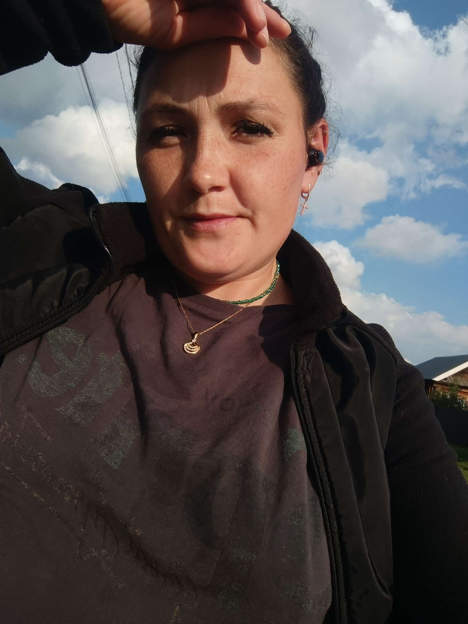 A medium shot photograph shows a woman outdoors against a sky with scattered clouds. The woman is looking towards the camera with her right hand raised to her forehead, shielding her eyes from the sun. She has dark hair pulled back and is wearing a black zip-up jacket over a dark t-shirt. A necklace with a gold pendant and a green beaded necklace are visible. She is also wearing a black earbud in her right ear and a small hoop earring with a cross charm. The background shows a glimpse of houses and a fence, suggesting a residential area. The lighting indicates it is daytime.