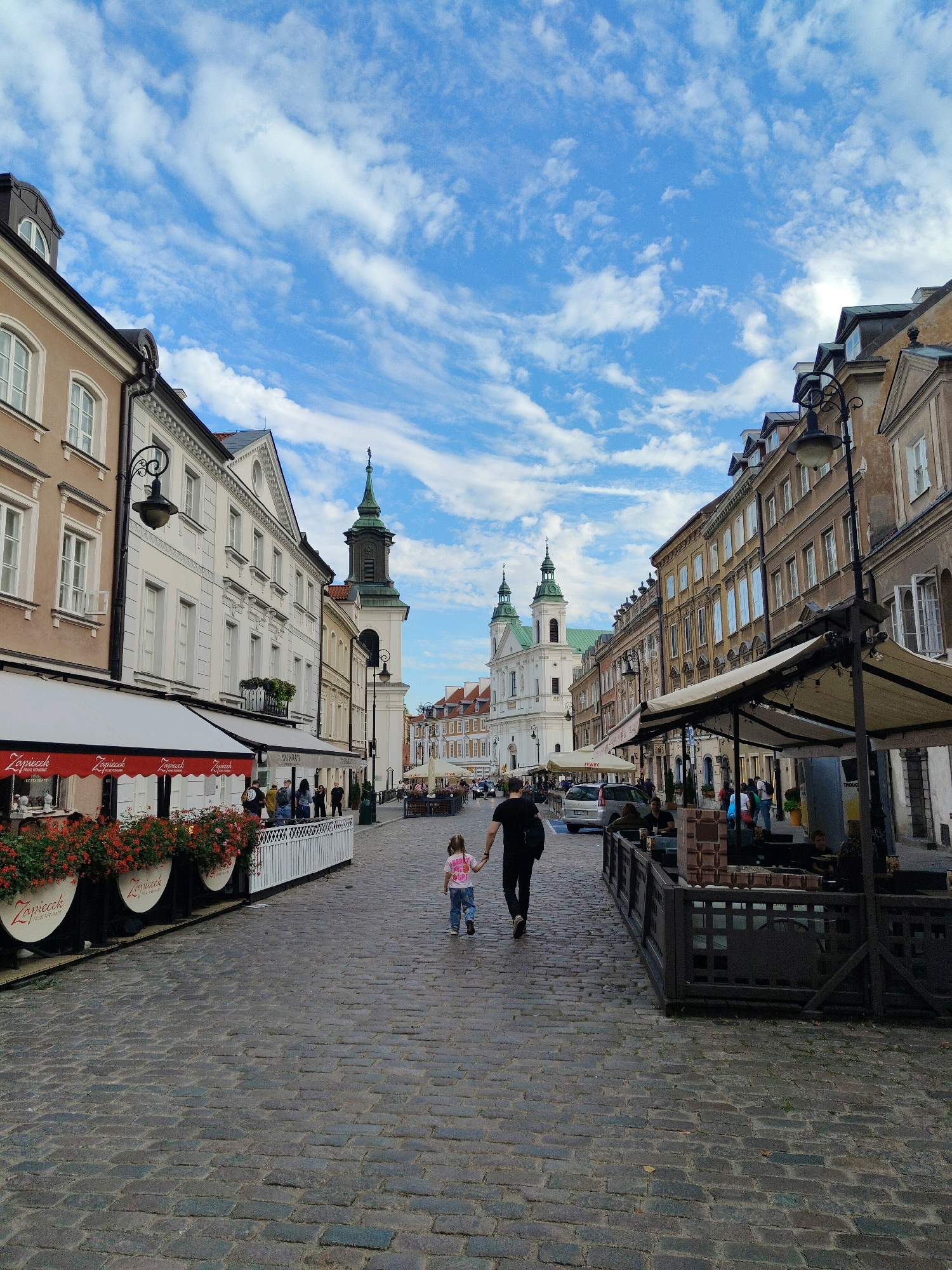 This is a vertical, eye-level, outdoor, daytime, full shot of a cobblestone street lined with historic buildings in Warsaw, Poland. A father and daughter are walking away from the camera toward the center of the frame. The father, wearing a black t-shirt and black pants, is holding the daughter's hand. The daughter, wearing a pink t-shirt and blue jeans, is walking to his left. The street is lined with cafes and restaurants, some with outdoor seating. In the background, a church with green spires is visible. The sky is blue with scattered clouds. The buildings on either side of the street are a mix of beige, white, and brown. The cobblestone street is well-maintained and shows signs of wear. The overall scene depicts a bustling urban environment with a blend of historic architecture and modern life.