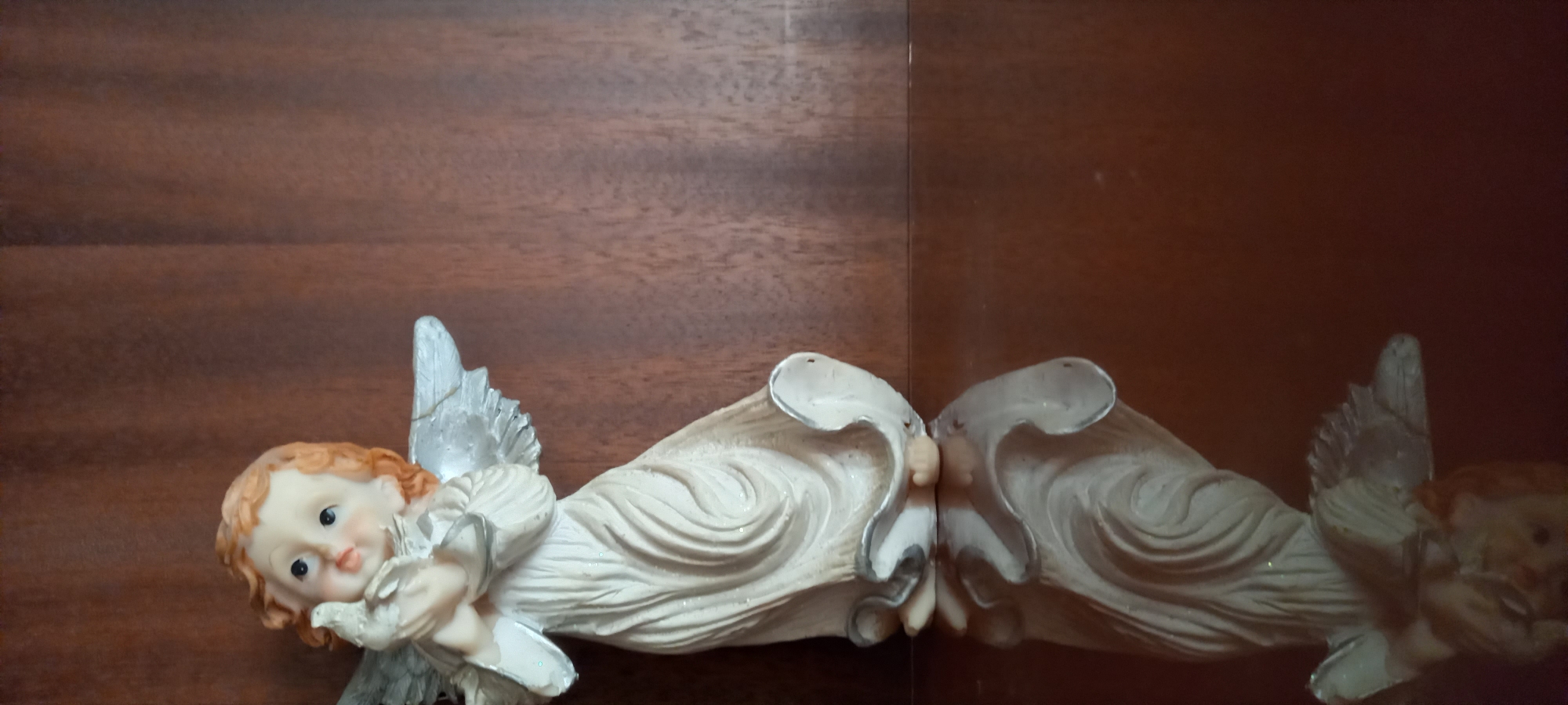 This image captures a close-up of a ceramic figurine depicting a cherubic angel, horizontally oriented in the frame. The angel has light reddish-brown, curly hair, fair skin, and delicate features, with a serene expression and small dark eyes. It wears a cream-colored gown, intricately sculpted with flowing drapery and subtle glittering details. White and silver-toned wings, showing textured lines, extend from its back. The angel is gently cradling a small white bird, likely a dove, in its hands.
To the immediate right of the figurine is its clear reflection, mirrored on a dark, glossy surface that creates a distinct vertical separation line in the center of the image. The reflection is slightly darker and less defined than the actual figurine. The background on the left side features a warm, reddish-brown wooden surface with visible horizontal grain, suggesting a piece of furniture or paneling. The area behind the reflection on the right is a darker, plain brown surface. The overall scene evokes a sense of peace and innocence. No text is visible in the image, which was taken in Kryvyi Rih, Ukraine.