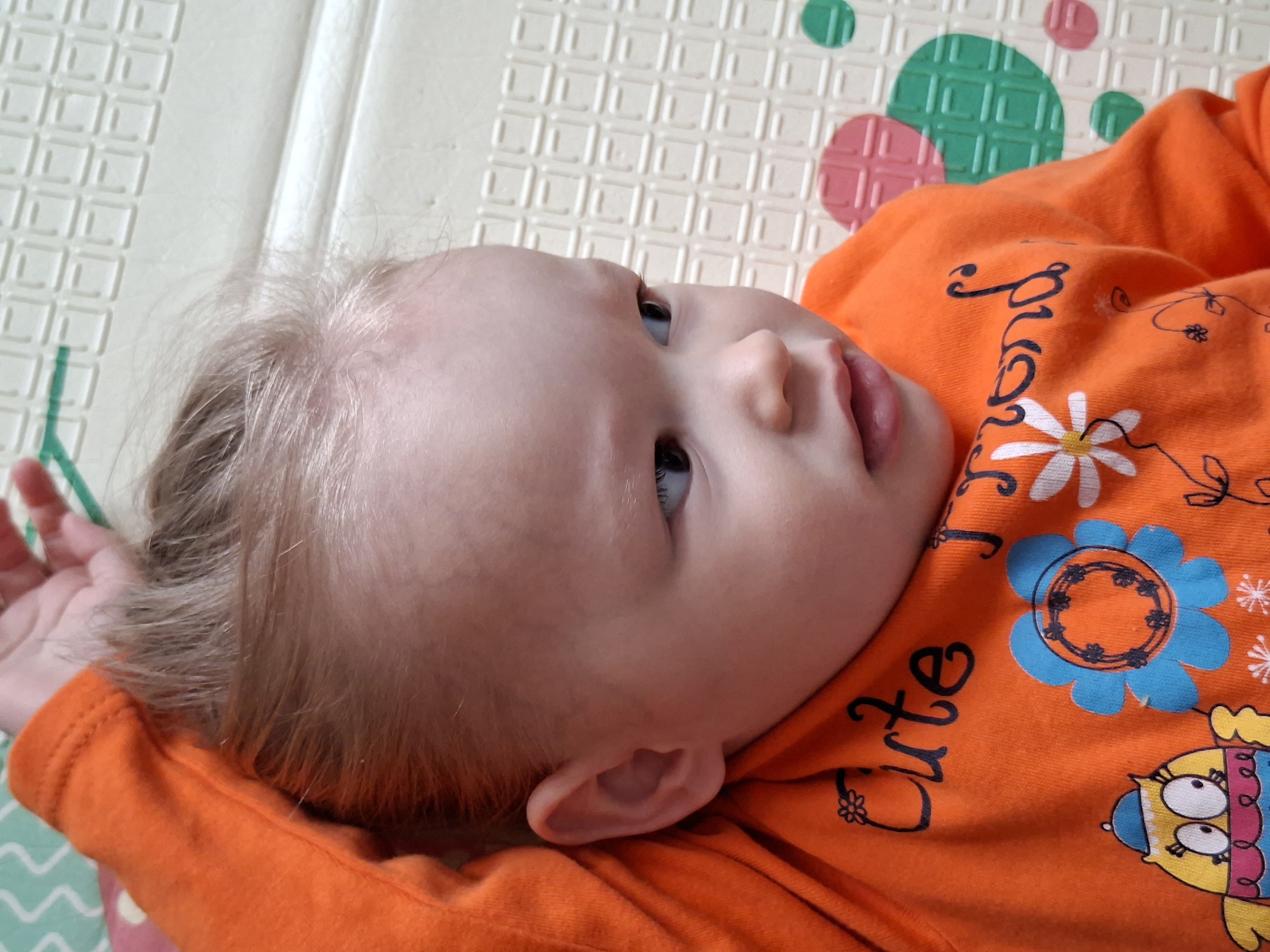 A close-up shot depicts a baby lying on a patterned mat. The baby has light hair and fair skin, and is wearing an orange shirt with floral and bird-like designs, along with the word "Cute" written on it. The mat beneath the baby features a grid pattern with colorful accents of green and pink circles and squares. The baby is looking towards the right of the frame with their mouth slightly open. The location appears to be indoors, based on the presence of the mat and the controlled lighting, but is not located in the city of Unknown, Unknown.