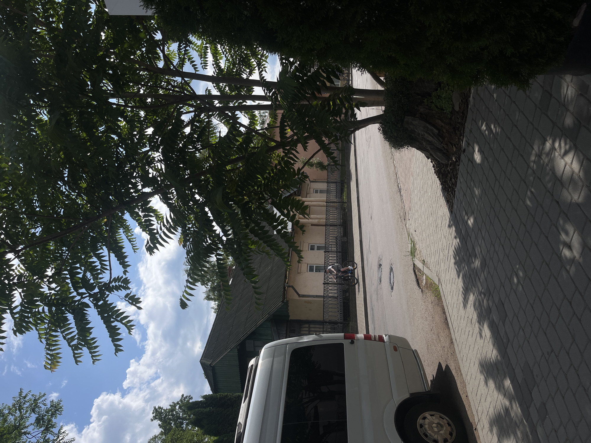 The image captures a street scene in Kosiv, Ukraine, on a sunny day with scattered clouds. In the foreground on the left, a silver van is parked parallel to a paved sidewalk. The van's rear section and wheel are visible. The sidewalk, made of grey interlocking pavers, extends across the bottom of the frame. Beyond the sidewalk, a street runs horizontally, with manhole covers visible on its surface.
On the right side of the street, a person is riding a bicycle. Further back, a pale yellow building with a dark-tiled roof and arched windows sits behind a decorative metal fence. Trees with green leaves are prominent throughout the scene, with a large tree partially obscuring the view of the sky and the building. The overall impression is a quiet, residential street on a pleasant day.