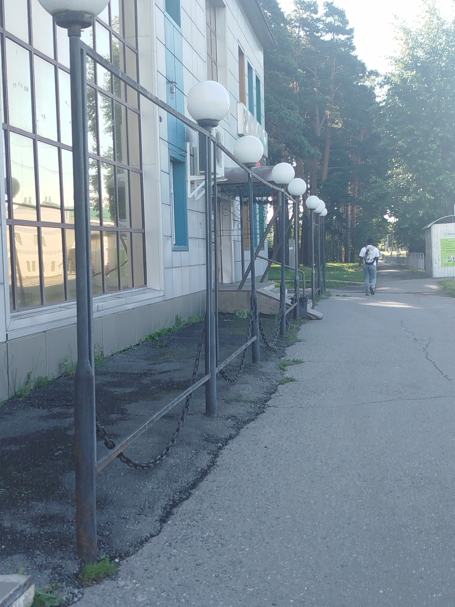 An outdoor, eye-level, full shot captures a person walking away from the camera on a paved path. The individual is wearing a white backpack and blue jeans. To the left of the path is a building with large, reflective windows. A row of spherical outdoor lights on metal posts lines the path, some with chains strung between them. Lush green trees are visible behind the building and beyond the path. The ground on either side of the path appears to be paved and somewhat cracked. The overall lighting suggests a daytime scene, possibly late afternoon, given the long shadows and the warm tones reflected in the windows.