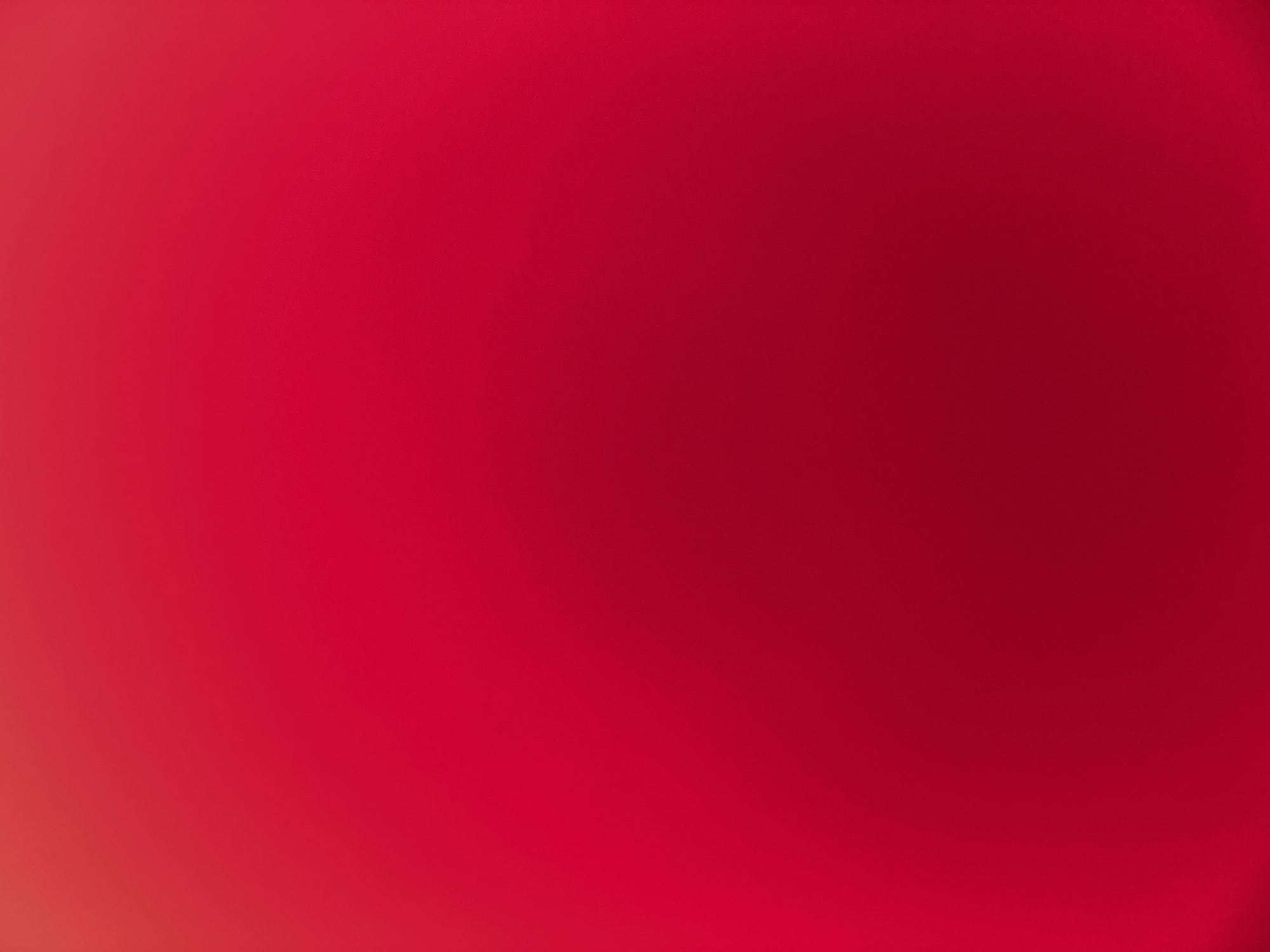 This media file is a full-frame, slightly out-of-focus abstract image dominated by a gradient of deep red. The color transitions subtly from a brighter, almost orangey-red on the bottom left to a richer, darker red towards the top and right sides. There are no discernible subjects, objects, scene elements, or specific activities depicted. The lighting appears soft and diffused, suggesting it might be an indoor setting or an overcast day, but there are no definitive cues to determine the time of day or weather. No location-specific details or emotional tones are evident due to the abstract nature of the image. There is no visible text within the frame. The image is a simple, monochromatic composition of red.