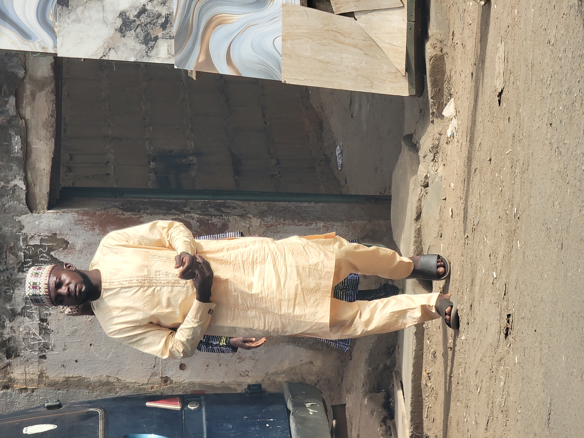 An adult Black male stands on a concrete sidewalk next to a street in the city of Maiburiji, Nigeria. He wears a light yellow, traditional, long-sleeved garment with matching pants and a patterned cap. His dark-skinned hands are clasped in front of him. He wears dark sandals on his feet. Behind him is a weathered building facade and some ceramic tiles leaning against it. Part of a dark-colored vehicle is visible in the lower left corner. The environment suggests an urban area with worn infrastructure.