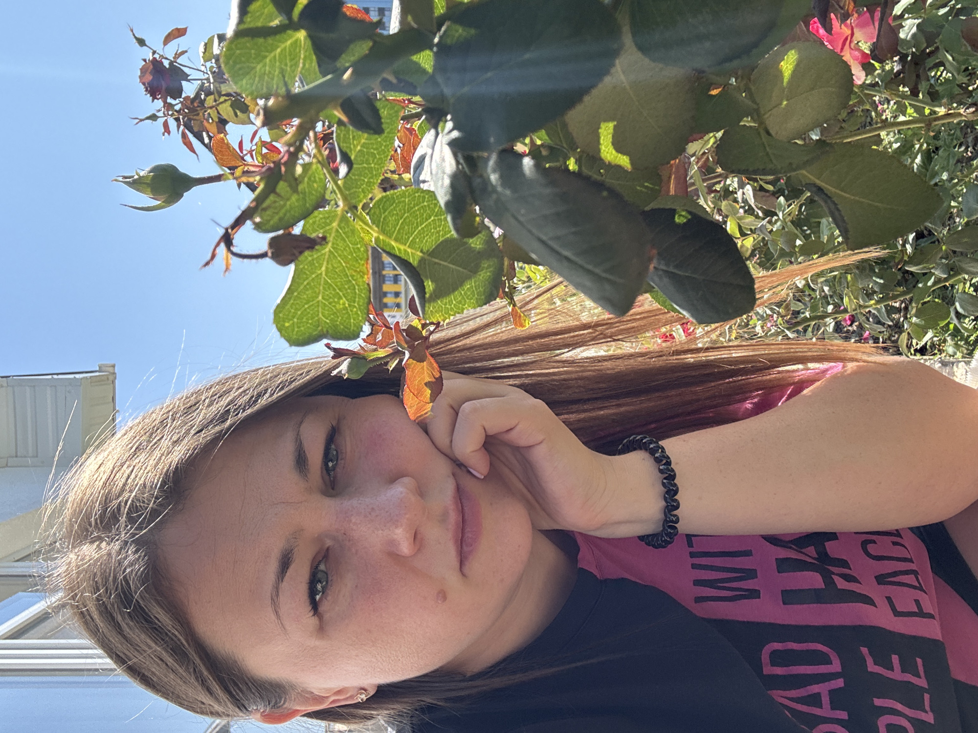 A medium shot portrait of a woman is captured outdoors in Uyutnoye, Ukraine, on a sunny day. The woman, with shoulder-length brown hair, is positioned on the left side of the frame, resting her chin on her right hand. She wears a black and pink t-shirt with white text that reads "BAD HABITS PEOPLE FACE". Her left earlobe sports a small earring. In the foreground and to the right of the woman, a rose bush with green leaves and a budding flower is visible, partially obscuring her. The background reveals a clear blue sky, the facade of a building with windows, and more of the rose garden. The lighting suggests it is daytime.