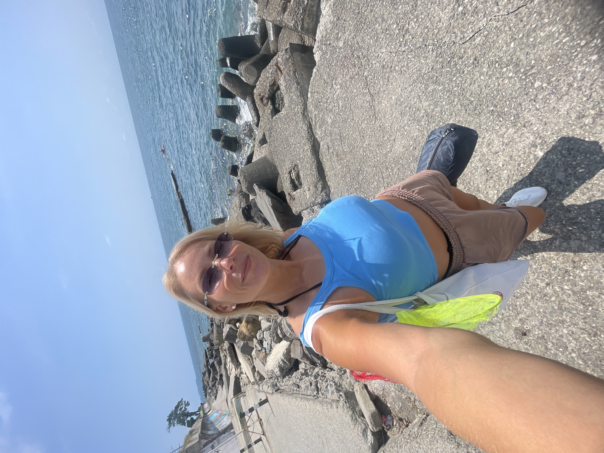A blonde woman with sunglasses takes a selfie on a sunny day. She is wearing a blue crop top and light brown shorts. She is carrying a white tote bag with a neon green mesh bag inside, and a black duffel bag. The woman is standing on a concrete path next to a rocky shoreline with the sea in the background. In the distance, a person is visible on a paddleboard in the water. The scene appears to be a coastal area in Sochi, Russia, indicated by the type of breakwaters and the style of the concrete promenade.