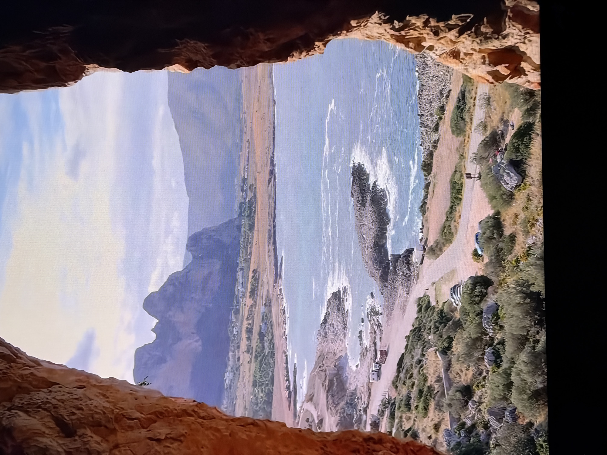The image shows a picturesque coastal landscape viewed from within a cave. The foreground is dominated by the textured, reddish-orange rock formations of the cave entrance, framing a panoramic view of the sea and surrounding terrain. The sea is a vibrant blue with white-capped waves breaking against a rocky shoreline. Beyond the coast, a dry, arid landscape stretches towards distant, hazy mountains. Several white camper vans are parked along a dirt road that follows the coastline. The sky is partly cloudy, with a soft, diffused light suggesting either early morning or late afternoon.
The location is NOT in the city of Chirchik, Uzbekistan. The scene depicts a Mediterranean coastline, likely in southern Europe, given the vegetation, rocky shores, and mountainous backdrop. The presence of camper vans and the overall topography are inconsistent with Uzbekistan's geography.