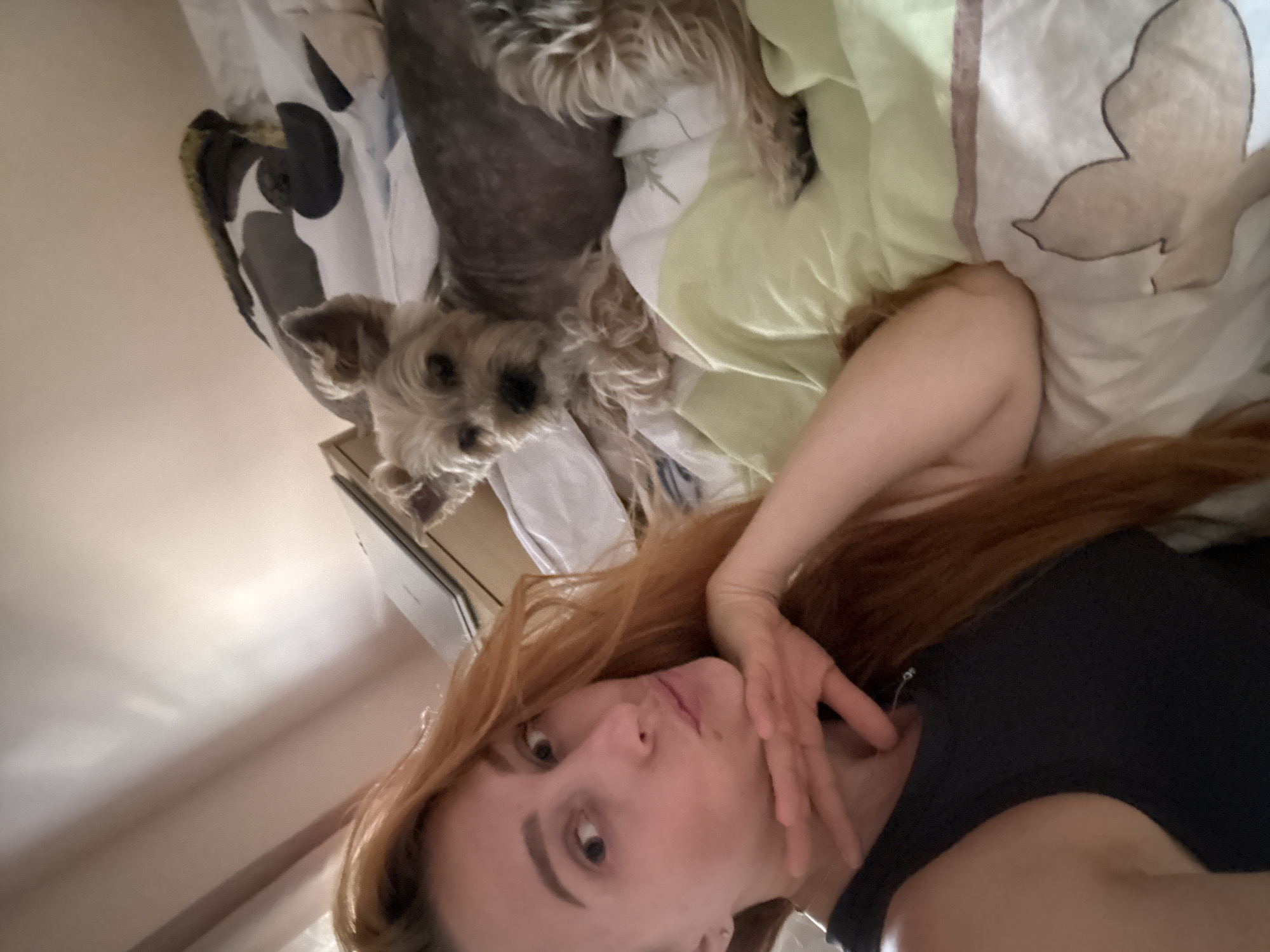 A woman with long, reddish-brown hair is lying on a bed, taking a selfie. She is wearing a dark tank top and has her left hand resting on her chin. Her lips are pursed, and she is looking towards the camera with a slightly raised eyebrow. To her right, a small, light-colored dog with scruffy fur is also lying on the bed, looking towards the camera. The bed is covered with patterned sheets and a light green duvet. In the background, a portion of a wall and what appears to be a laptop are visible. The lighting suggests an indoor setting, likely a bedroom. The presence of a bed and the relaxed posture of both the woman and the dog indicate a private, domestic environment. No specific landmarks or features in the image definitively place the scene in Budva, Montenegro.