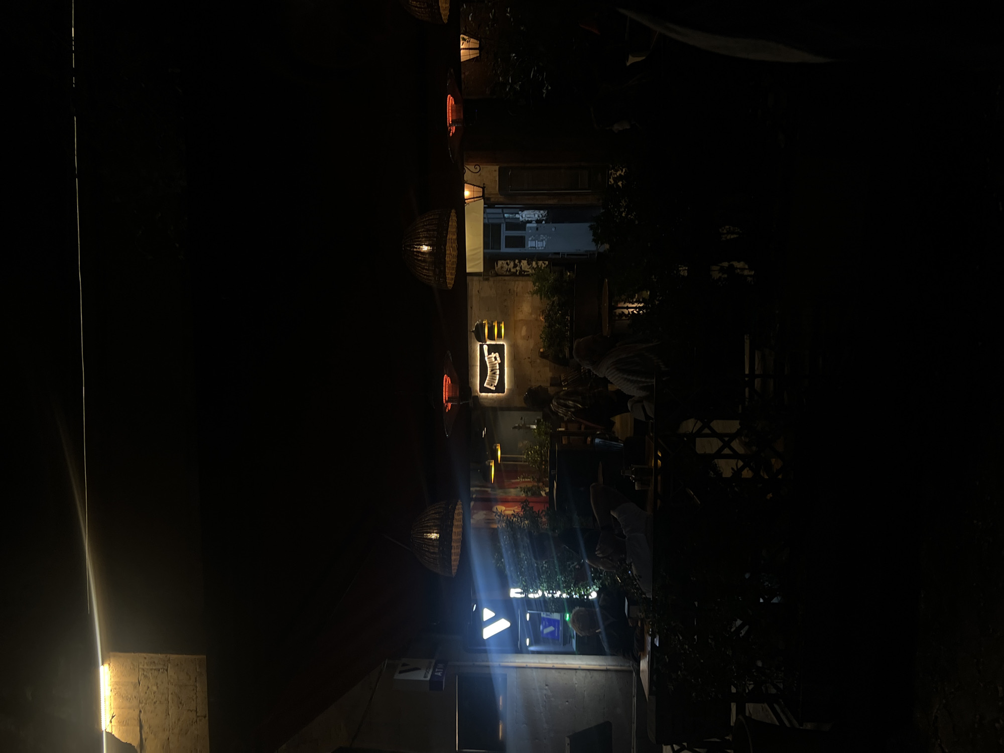 The image depicts an outdoor area, likely in Unknown, Unknown, illuminated by various artificial lights at night. The location appears to be a restaurant or cafe, indicated by the presence of tables and chairs occupied by people. Several decorative lamps hang overhead, casting warm light downwards. A sign with the name of the establishment is visible on the wall. The overall atmosphere is dimly lit and suggests a relaxed, urban setting. Vegetation, including trees or bushes, is present in the background, adding to the ambiance. The image also includes a sign that says “ATM” and an arrow.