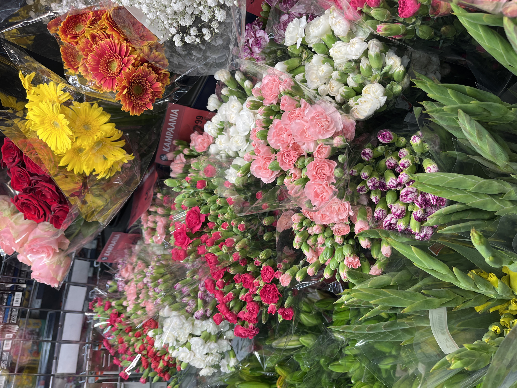 The image shows a close-up, eye-level view of a colorful assortment of flowers displayed in clear plastic packaging, likely in a supermarket flower section. In the foreground, a large bunch of gladiolus with green stalks and yellow buds is prominent. Behind and to the right, various carnations are visible in shades of pink, white, and red, some in full bloom and others as buds. To the upper right, bouquets of yellow and orange-red gerbera daisies are partially visible, along with a bunch of small white flowers, possibly baby's breath. In the upper left, pink and red roses are also displayed. A sign with "KAMPANJA" and a price is visible amongst the flowers, indicating a sale. The background is blurred but suggests shelving and other products, consistent with a retail environment. The location is specified as Kohtla-Järve linn, Estonia, but the image itself does not contain explicit visual cues to confirm this specific location, relying solely on the provided metadata. There are no people visible in the image.