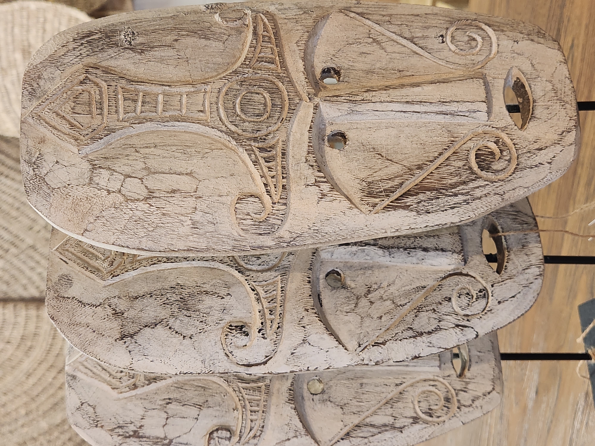 The media file displays a close-up, overhead view of three carved wooden objects stacked on top of each other. The objects are oval-shaped and feature intricate, abstract carvings with swirling patterns and geometric shapes. The wood appears weathered and has a light, distressed finish, with darker areas accentuating the carved details. Each object has two circular holes, suggesting they might be intended for hanging or for some other functional purpose. The background is softly blurred, hinting at a rustic or artisanal setting, possibly a market stall or workshop, with woven textures and wooden elements visible. The lighting suggests it is daytime, with natural light illuminating the carved surfaces and highlighting the textures of the wood. No text is visible in the image. The overall impression is one of handcrafted artistry and a natural, earthy aesthetic. The setting appears to be Maitland, United States of America.
