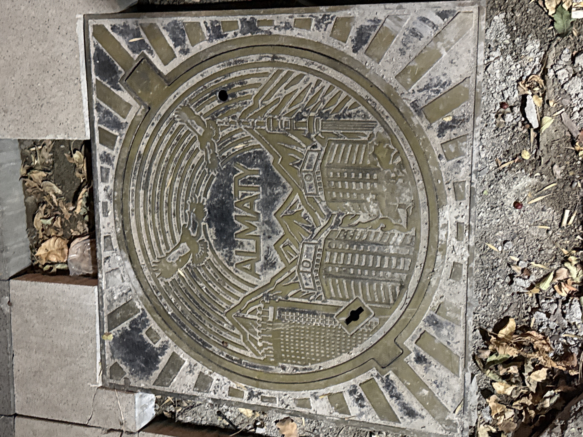 A circular, metallic manhole cover is the central focus of the image, resting on a paved surface that appears to be a sidewalk or street. The manhole cover is intricately designed with raised relief patterns. At the center, the word "ALMATY" is prominently displayed in capital letters. Surrounding the text are depictions of mountains, a tall tower, and several multi-story buildings, suggesting a cityscape. Two birds are etched into the upper portion of the design. The outer rim of the manhole cover features a geometric pattern of alternating dark and light rectangular shapes. The manhole cover is weathered and shows signs of wear, with some dirt and debris accumulated on its surface and around its edges. The surrounding environment consists of cracked concrete paving stones and patches of loose gravel and soil. Fallen leaves are scattered on the ground, indicating an outdoor setting, possibly in a park or along a street with trees. The lighting suggests it may be dusk or nighttime, given the somewhat dim illumination and the shadows cast.
