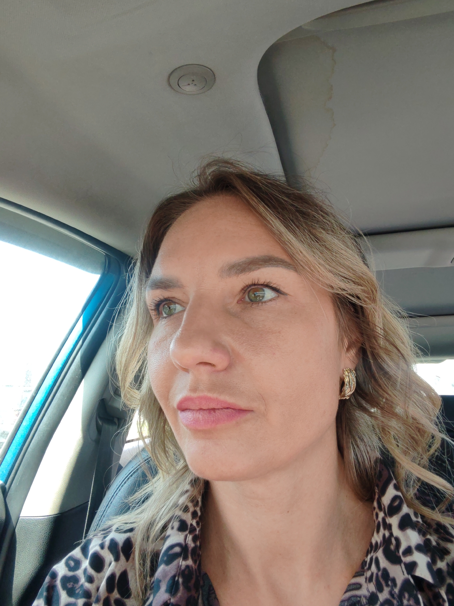 A close-up, eye-level shot features a woman seated in a car, her head and upper torso filling the frame. She is looking to her left (our left), with her gaze directed outwards from the vehicle. She has light brown to blonde, wavy hair and distinctive green eyes. Her face shows minimal makeup, with soft pink lipstick. She wears gold-colored, textured hoop earrings and a brown and black leopard print top.
The setting is inside a car, evidenced by the light grey fabric headliner of the car ceiling, which features a circular vent or light fixture. A prominent brownish-yellow stain is visible on the ceiling, particularly near what appears to be a sunroof or overhead console. Bright, diffused natural light streams in from the driver's side window, indicating it is daytime. A segment of a blue interior car trim is visible along the window's edge, and a black car seat is partially seen behind her shoulder. The woman appears to be observing her surroundings or in thought. The overall scene suggests a casual moment during a daytime car ride in Petropavlovsk-Kamchatsky, Russia. There is no visible text in the image.