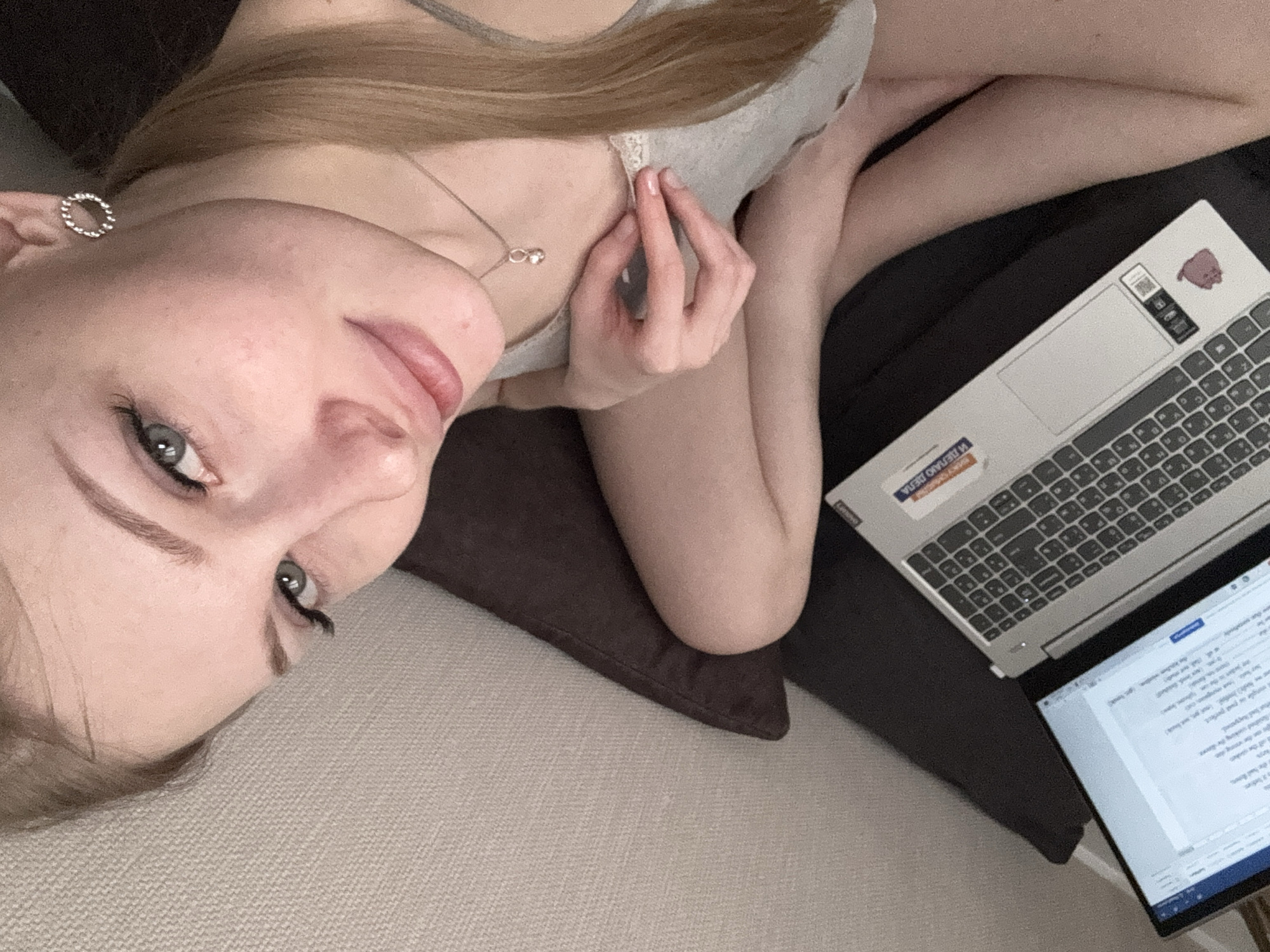 A female individual is depicted in a high-angle, close-up indoor shot, located in Perm, Russia. She has light skin, light blonde hair pulled into a ponytail, blue-green eyes with dark eyeliner, and pink lips displaying a faint smile. She is wearing a light grey tank top with white lace trim, a silver-colored chain with a small heart-shaped pendant, and a silver-colored circular textured stud earring in her right ear. Her right hand, with manicured fingernails, rests on her chest near her collarbone. A silver-colored Lenovo laptop, featuring a Cyrillic keyboard and multiple stickers including one partially legible as "ДЕЛАНО", is positioned to the lower left. The laptop's screen displays a word processing document. The individual's bent knees are visible in the lower center. A dark brown cushion supports her, and a light-colored, textured surface, likely a wall or upholstered furniture, forms the background. She looks directly at the camera.