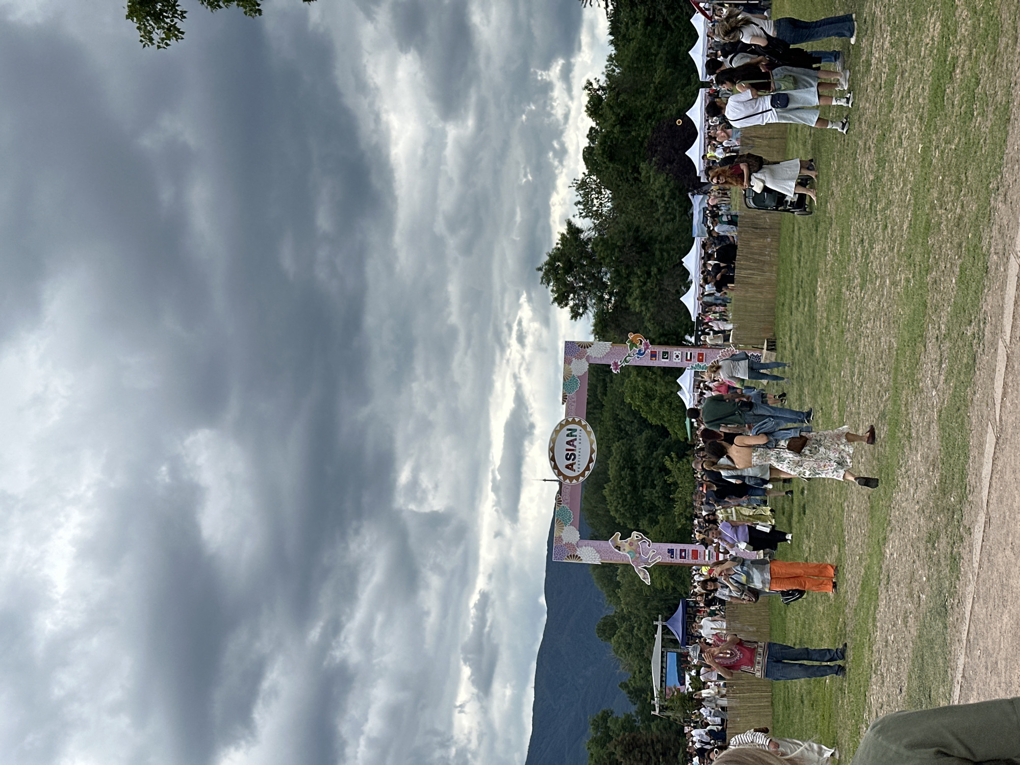 This is an outdoor festival scene with a large crowd of people milling about under a cloudy sky. In the foreground, there is a dirt path and a grassy field. In the midground, an archway with the text "ASIAN FESTIVAL SOFIA" and decorative elements like flowers and a horse is set up. Several white tents are visible behind the archway, suggesting vendor stalls or information booths. A stage with a screen can be seen on the left side of the frame. In the background, lush green trees and mountains are present. Numerous individuals are walking, standing, and some appear to be taking photos. The overall atmosphere suggests a public event or celebration taking place in a park-like setting within Sofia, Bulgaria.