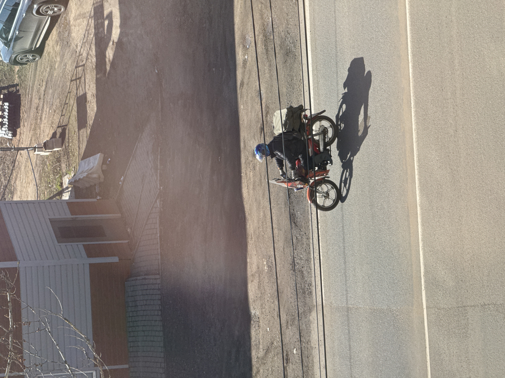 Captured from above, the image shows a man on a moped traveling along a road. The man is wearing a helmet and dark clothing, and the moped is carrying a bag. The road is bordered by a sidewalk and utility poles. To the left, there's a house and vegetation. In the upper left corner, a portion of a car is visible. The scene appears to be in daylight. The location is identified as Ново-Никольское, Russia.
