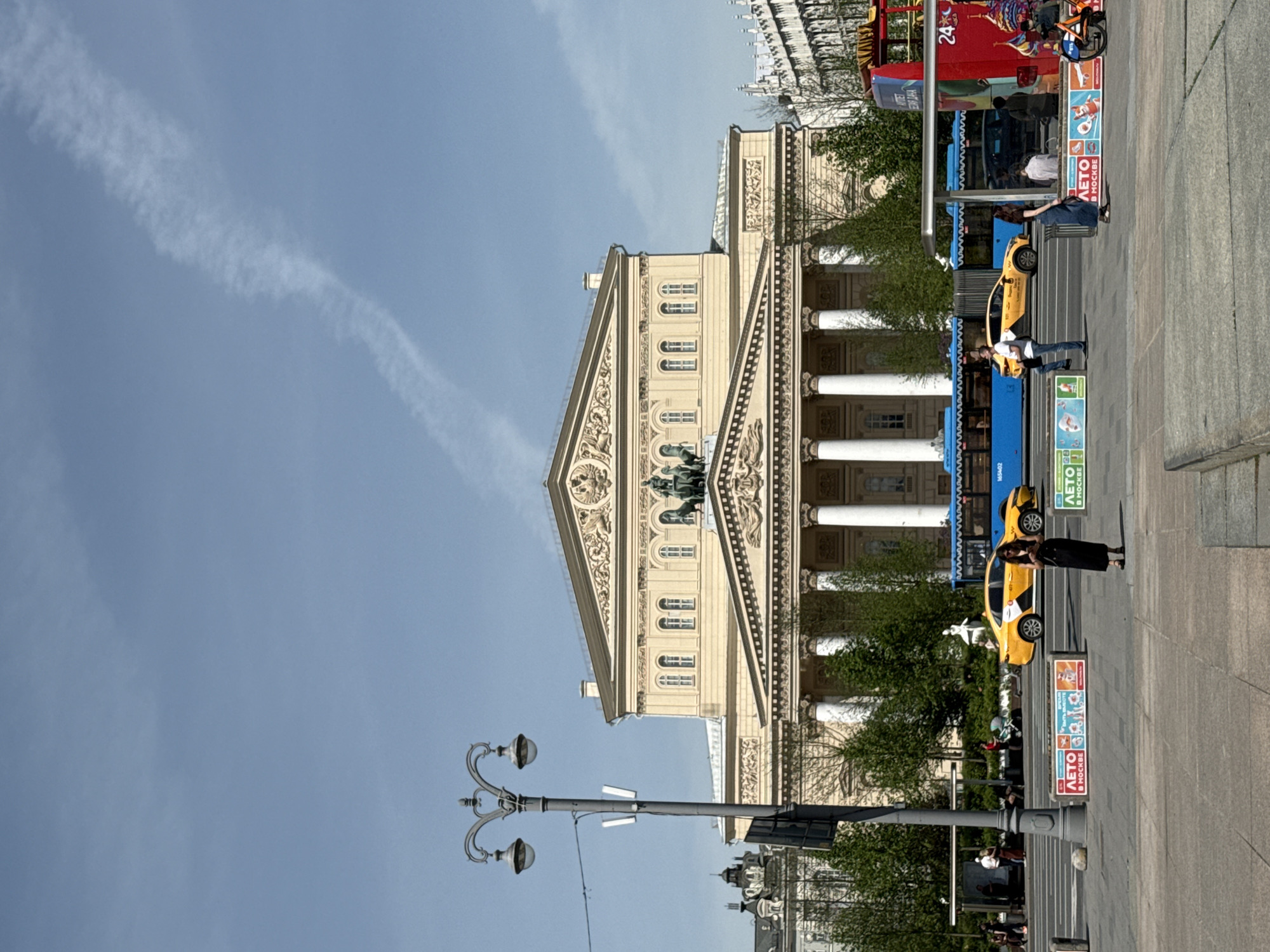 The image shows the Bolshoi Theatre in Moscow, Russia. The building's facade is visible, including its columns, statues, and decorative elements. A blue and red tour bus and yellow taxi are on the street beside the theatre. A few people are standing and walking around. A light pole is on the left. The sky is clear with one cloud.