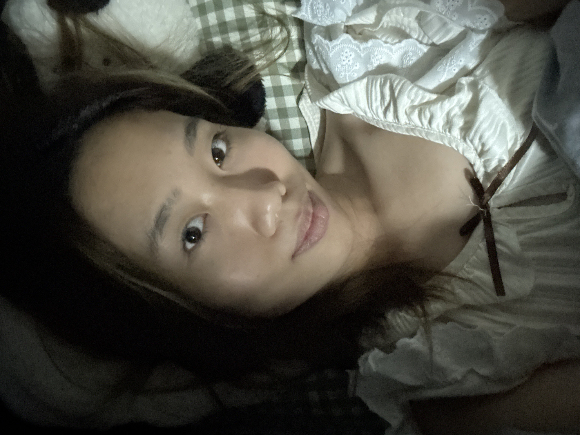 A woman lies on a green-and-white checkered surface, possibly a bed, wearing a white top with lace and a brown tie at the neckline. Her dark hair is visible on one side, with a black headband. The woman faces the camera, slightly smiling. The lighting is dim. There is no indication of a location in Nan'an District, China.