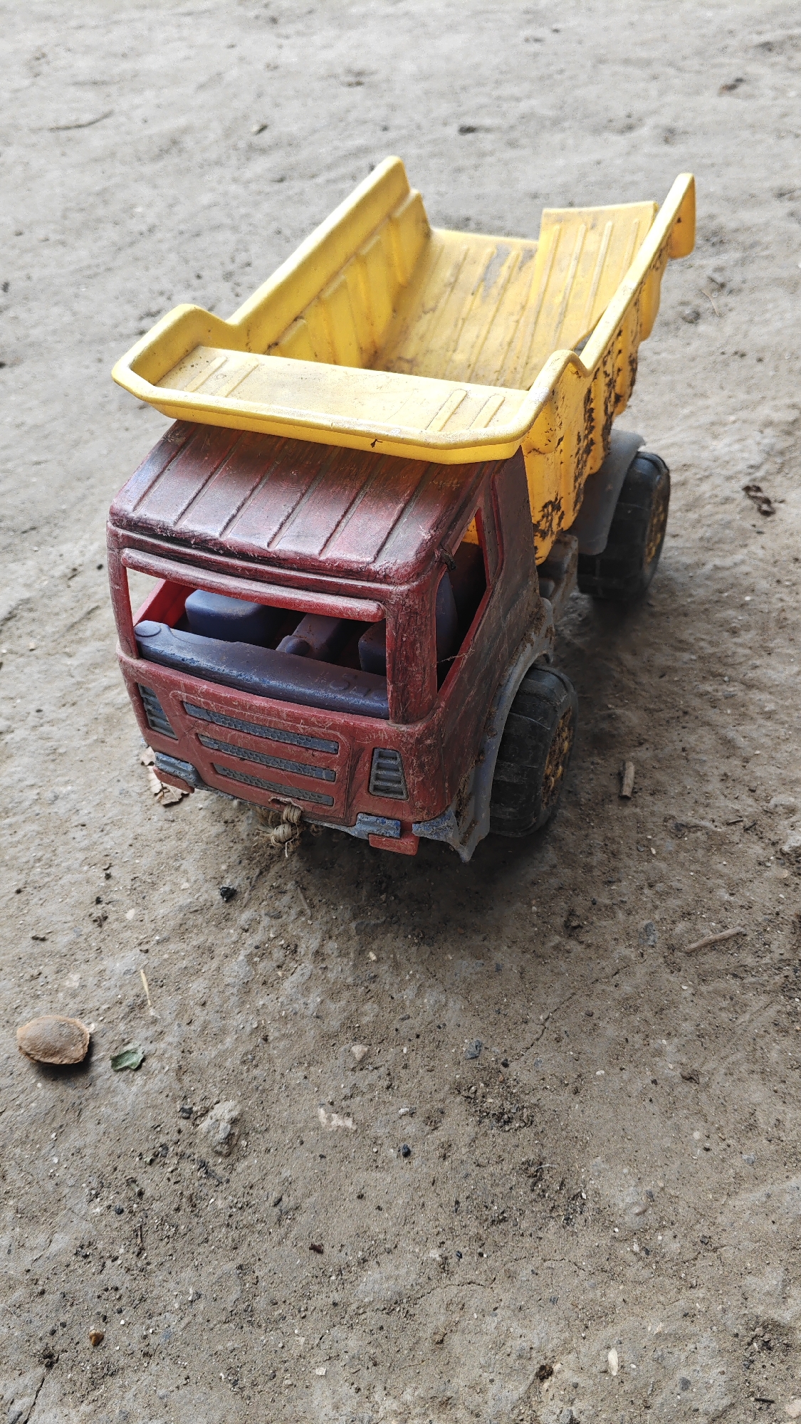 An overhead shot of a weathered, red and yellow toy dump truck sits on a dirt surface. The truck's cab is red with blue seats visible through the windows. The truck bed is yellow and appears to be stained and dirty. The tires are black and appear to be made of rubber. The ground is composed of light brown dirt with small pebbles and debris scattered across it. In the lower left corner of the image, there is a small, brown, oval-shaped object, possibly a seed or a small stone, and a single green leaf. The background is out of focus but appears to be more of the same dirt surface.