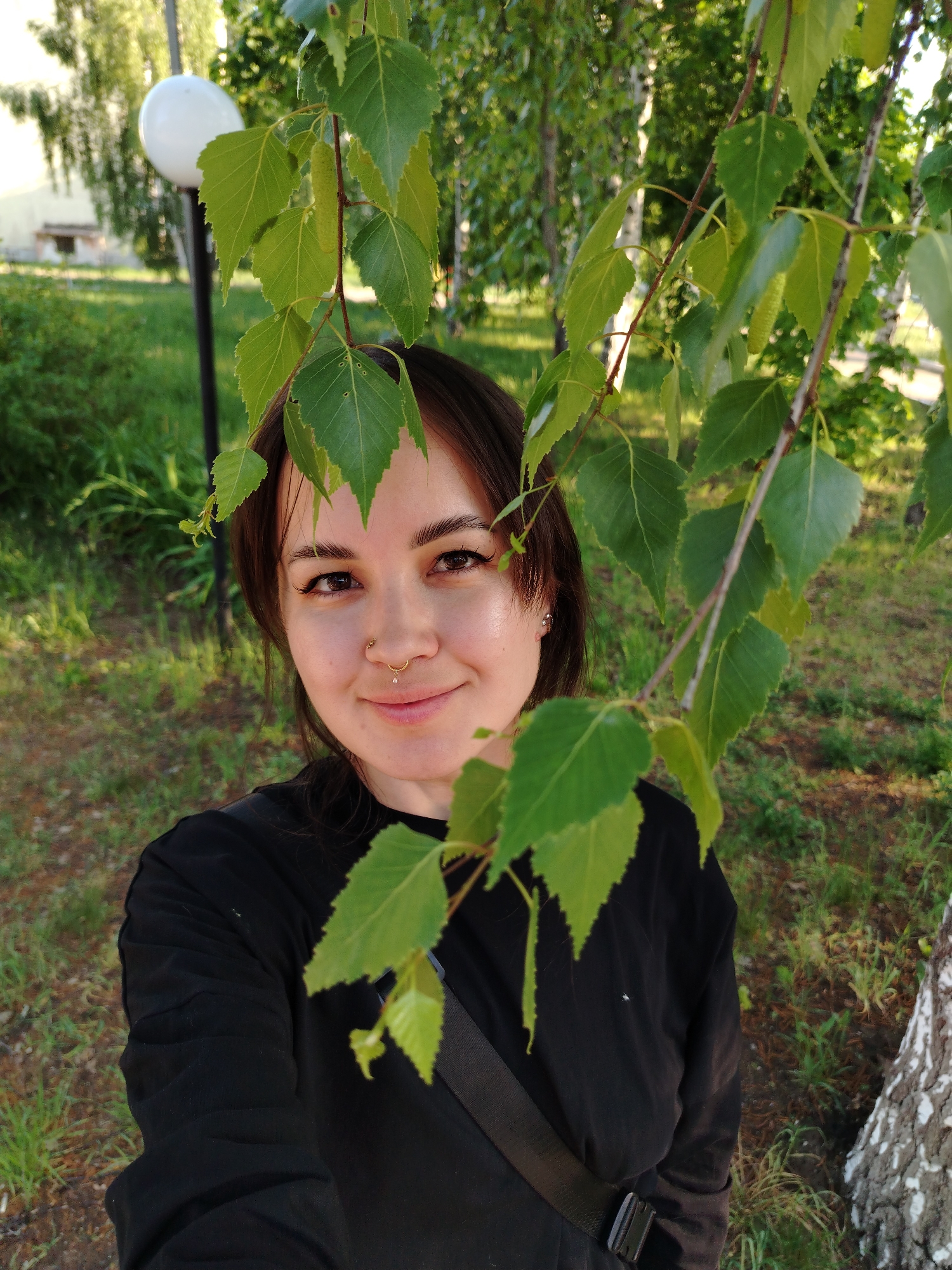 A young woman with dark hair and brown eyes stands outdoors amidst foliage. She has a nose ring and wears a black long-sleeved shirt with a strap visible on her shoulder. Green leaves from a birch tree are prominent in the foreground, partially obscuring her. The background shows a grassy area, trees, a building, and a lamppost with a globe-shaped light. The location is Noviy, Russia.