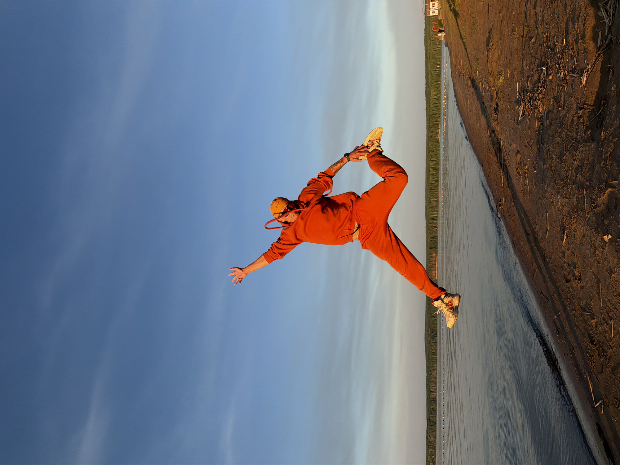 The image displays a person performing a complex balancing pose, similar to a variation of Natarajasana (Lord of the Dance pose), on a sandy or muddy riverbank. The subject, clad in a vibrant orange tracksuit, a yellow beanie or hood, and light-colored sneakers, stands on one leg. Their other leg is bent backward, with the foot held by both hands behind their back, creating an elegant arch in their body. One arm is extended forward and slightly upward, with an open palm, aiding in balance. A watch is visible on their left wrist.
The setting is outdoors, alongside a body of water, likely a river or large lake, with a dark, earthy bank that shows signs of sand and small debris. A line of buoys or markers stretches across the calm water. In the background, beyond the water, a dense line of green trees or forest is visible, along with a few small buildings on the distant shore.
The sky is clear and prominent, transitioning from a lighter, almost white-blue near the horizon to a deeper blue overhead, with faint wisps of clouds. The warm, golden light suggests late afternoon or early evening, possibly during sunset. The scene evokes a sense of tranquility and focused activity, showcasing the person's balance and flexibility against a natural backdrop, presumably in or near Chaikovsky, Russia. There is no visible text in the image.