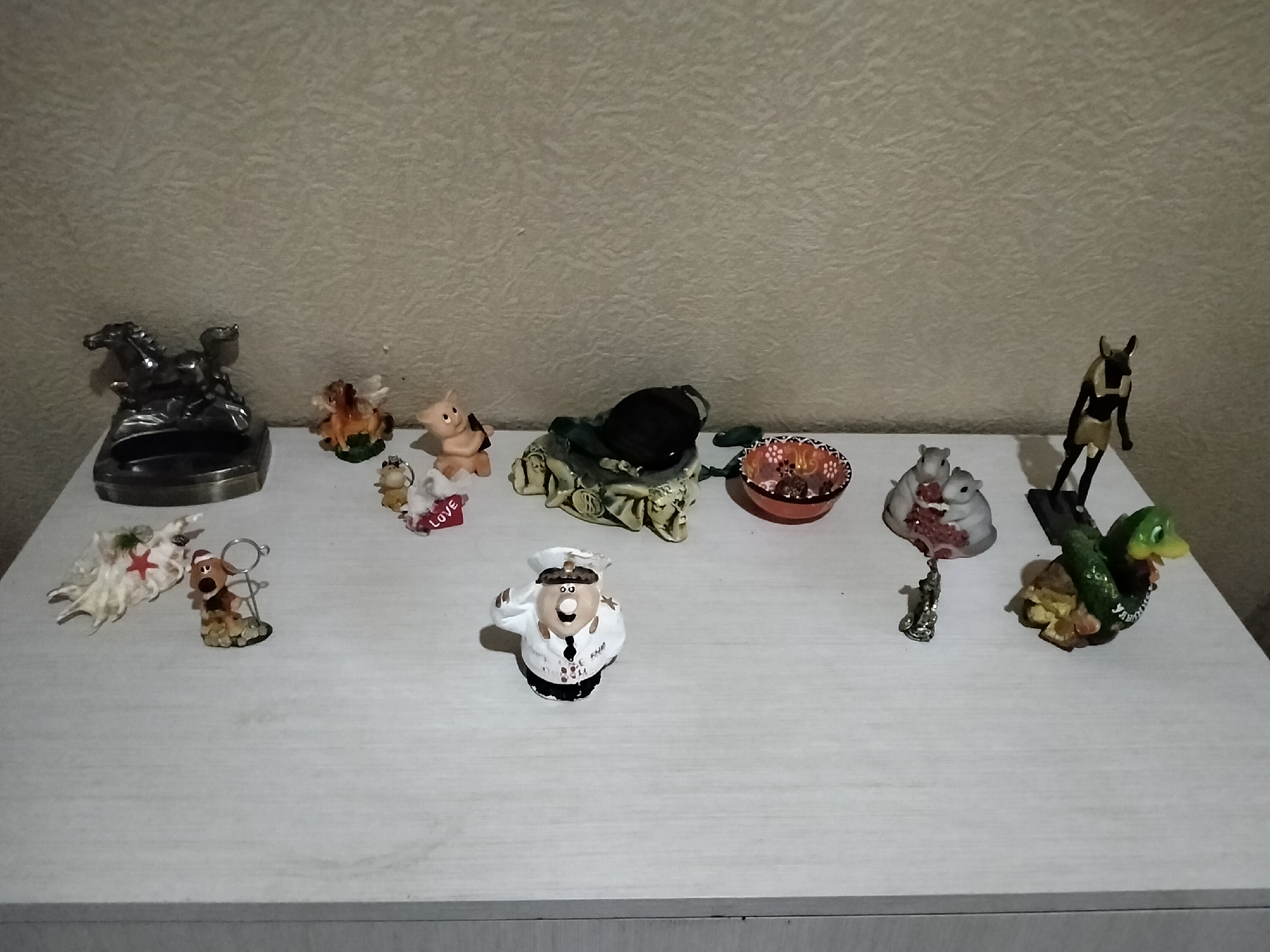 The image shows a collection of small decorative figurines displayed on a white, wooden surface, possibly a shelf or a dresser. The background is a textured, light-colored wall.
Starting from the left, there is a metallic sculpture of two horses, one rearing and the other in a galloping stance. To its right, a small, winged figurine with brown hair stands on a green base. Next to that is a pink pig figurine. Further to the right, a light-colored figurine, possibly a dog wearing a Santa hat and holding a small stick, is positioned on a speckled base. Next to the dog figurine is a white, fluffy object with a red star in the center, resembling a snowflake or a stylized creature.
Moving towards the center, there is a dark, rounded object, perhaps a decorative stone or a paperweight, placed on what appears to be a green, cloth-like material adorned with dollar signs. To the right of this, a small, orange and red patterned bowl sits on the surface.
Further to the right, there are two small figurines of squirrels. One is light gray and the other is a lighter shade of gray. A tiny silver figurine hangs from the squirrel figurines, resembling a person in a running pose.
To the right of the squirrels is a dark figurine of a figure with a dog-like head and humanoid body, standing upright. Next to it, a green ceramic duck figurine is depicted with its mouth open and wings spread, resting on a cluster of yellow and brown shapes, possibly representing flowers or leaves.
In the foreground, slightly off-center, is a figurine of a person dressed as a chef, wearing a white uniform, a white hat, and a black collar. The figurine has a round face with visible eyes, a nose, and a mouth, and is saluting with its right hand. The chef figurine has text on its uniform, but it is difficult to decipher.
The overall arrangement suggests a personal collection of decorative items. The image does not contain any recognizable landmarks or clues to indicate its location within Nizhnedonskoy, Russia.