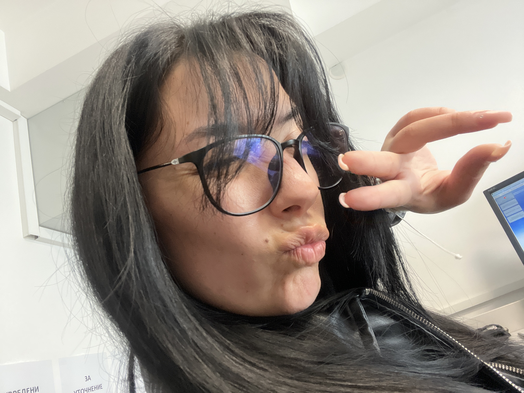 A woman with long, dark hair and wearing black-rimmed glasses is making a kissing face at the camera. She is holding her right hand up, fingers partially curled. She is wearing a black jacket. A headset with a white cord is present near her hand and ear. In the background, there is a computer monitor and a wall with two pieces of paper, one labeled "ЗА УТОЧНЕНИЕ". Based on the text on the papers, the location is likely Saedinenie, Bulgaria. The lighting appears to be artificial.
