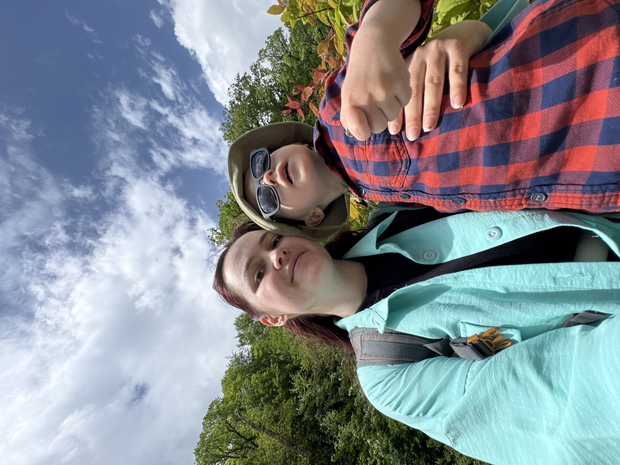 The image shows a woman and a young child taking a selfie outdoors. The woman is in the foreground, wearing a light blue button-down shirt and a backpack. The child is behind her, wearing a red and blue plaid shirt, a sun hat, and sunglasses. They are both smiling. The background consists of trees and a blue sky with white clouds. The photo appears to be taken in a natural setting, possibly a park or hiking trail. It is not possible to confirm that the photo was taken in the city of Estosadok, Russia, based on the visual information in the image alone.
