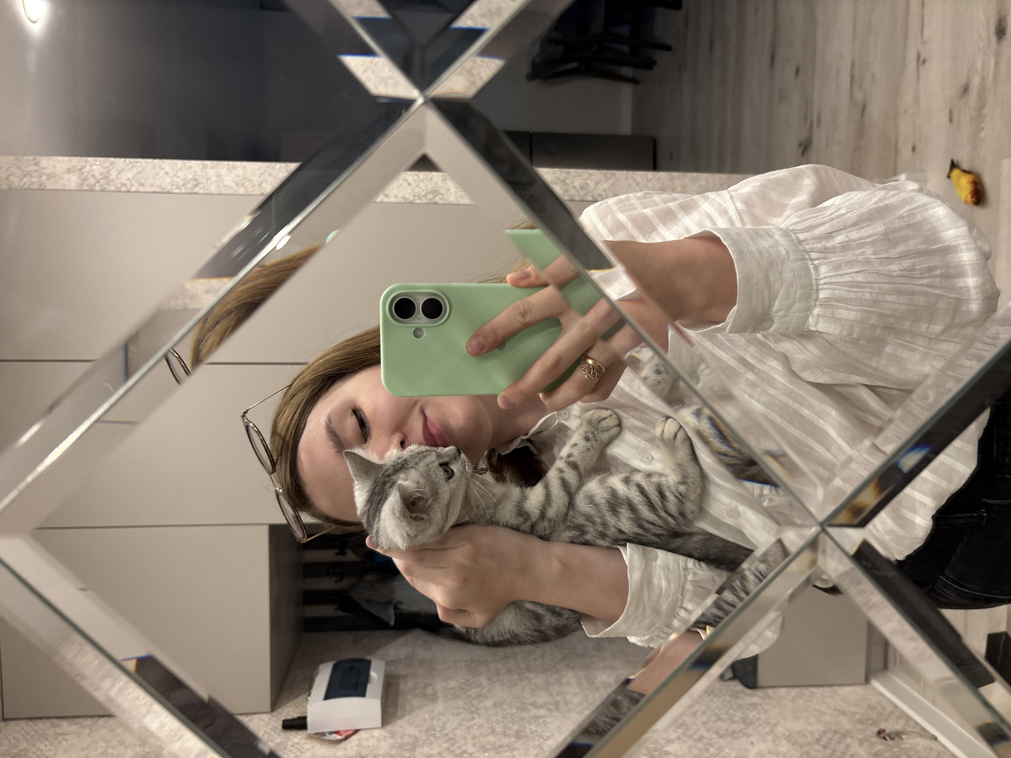 A young woman with light brown hair, wearing a white blouse and eyeglasses perched on her head, is taking a mirror selfie. She is holding a small, grey tabby kitten with stripes against her chest. The woman is holding her smartphone, which has a light green case, in her right hand, and her left arm is supporting the kitten. She is smiling gently, and the kitten appears to be looking to the side. The background of the image, seen in the mirror, shows an interior space with textured walls, a white cabinet, and a floor that appears to be wood. The mirror itself has a decorative, faceted frame. The image was taken in an apartment in Izhevsk, Russia.