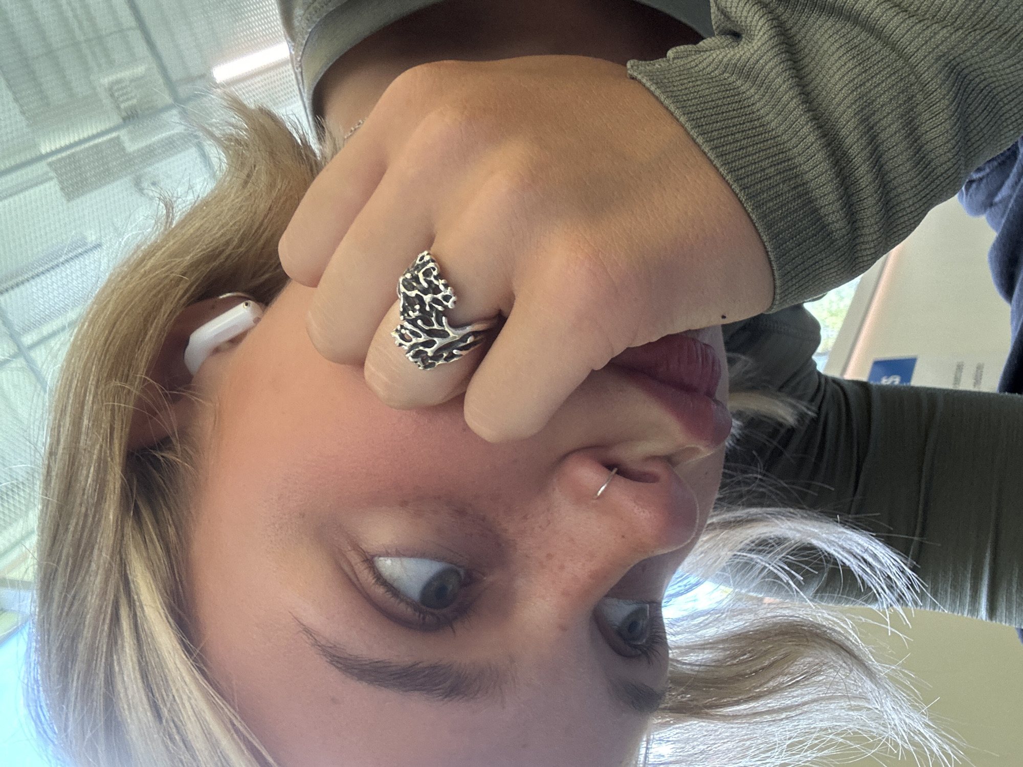 The image is a close-up, angled selfie of a young woman with blonde hair, fair skin, and light eyes. She is wearing a grey, ribbed long-sleeved shirt and has a silver ring on her right index finger, shaped like a coral or tree branch. A white wireless earbud is visible in her left ear. She has a small silver hoop piercing in her nose. Her right hand is resting on her cheek, and her gaze is directed downwards and to the left. The background is out of focus and appears to be an indoor environment with a ceiling grid and lighting fixtures. There is no discernible information in the image to suggest it was taken in Whistler, Canada.