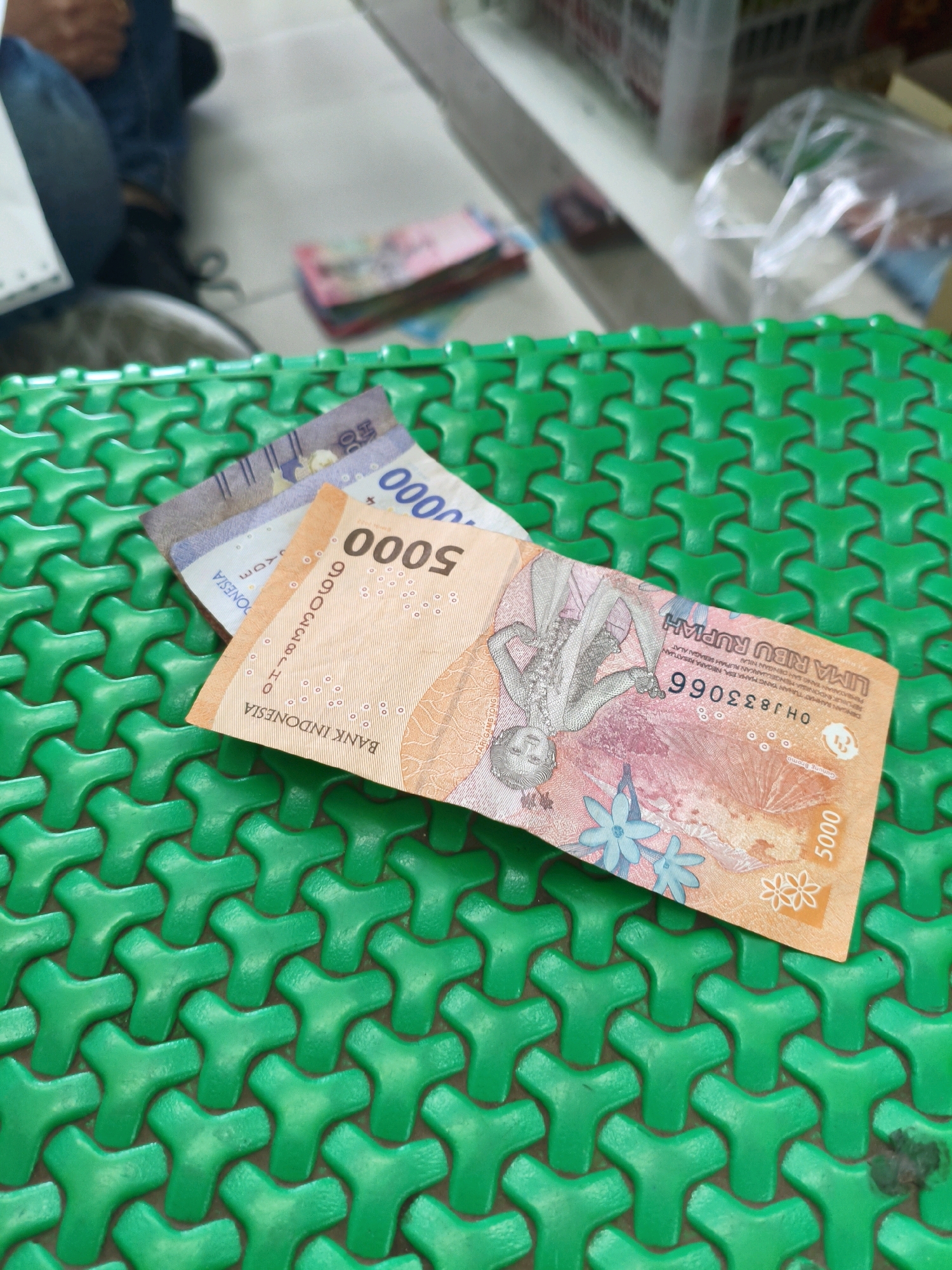 A close-up, eye-level shot shows two Indonesian Rupiah banknotes, a 5000 denomination and a portion of another, resting on a bright green, textured surface with a repeating tri-pronged pattern. The 5000 Rupiah note is predominantly orange and features an engraved image of a male figure and floral designs. Text on the banknote includes "BANK INDONESIA" and "LIMA RIBU RUPIAH." The background is blurred, showing a floor and people seated, suggesting an indoor setting possibly in Sumberejo Kulon, Indonesia.