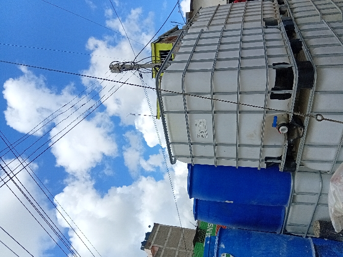 The image shows a large white rectangular water tank with a metal frame. The tank is sitting on a pallet on a concrete surface, with the front side partially covered by blue plastic barrels. The tank is situated in front of a building with a green roof and a utility pole with electrical wires running overhead. The sky is a bright blue with white clouds. The coordinates (-1.396, 36.942) place the scene in the town of Mlolongo, Machakos County, Kenya. The scene appears to be a typical residential or industrial area, with evidence of construction and infrastructure development.