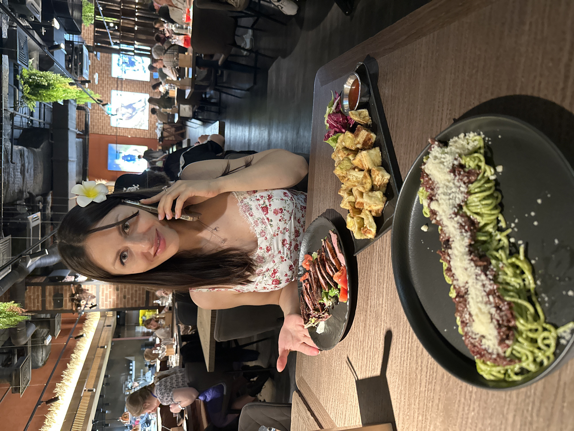 The photograph shows a woman sitting at a table in a restaurant. She is fair-skinned with long brown hair, and is wearing a floral print dress. She is holding a cell phone to her ear and has a white flower in her hair.
On the table in front of her there are three plates of food. One plate has what appears to be sliced steak and tomato, another contains fried food with a dipping sauce and lettuce, and the third contains a dish of green spaghetti topped with a dark topping and grated cheese.
The restaurant appears to be dimly lit and has a brick wall in the background. There are other people dining in the restaurant. Television screens on the wall display images, and indoor plants hang from the ceiling. Based on the style and interior design, the photograph appears to have been taken in Saint Petersburg, Russia.