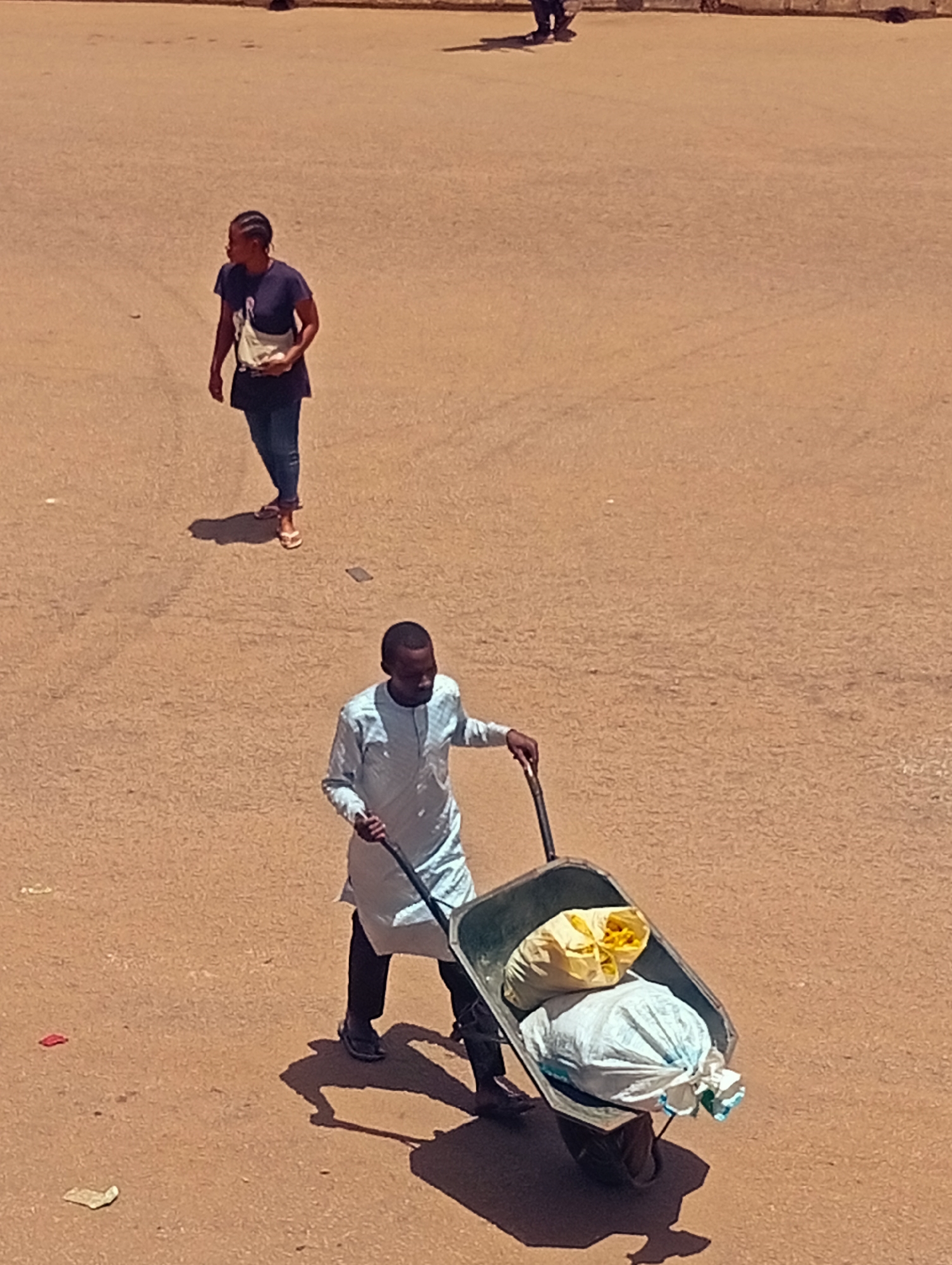 The image shows an open-air scene in the city of Unknown, Unknown. A man is seen pushing a wheelbarrow containing what appear to be bagged materials. A woman is walking at a distance away from the man. The ground is a flat, barren surface.