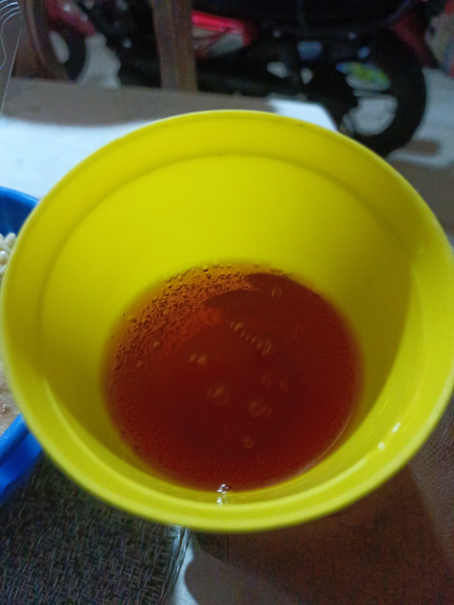 A top-down shot shows a yellow plastic cup partially filled with a reddish-brown liquid. Small bubbles are visible throughout the liquid, especially around the edges where the liquid meets the cup. A small portion of a blue plastic container is visible in the lower left corner of the frame, alongside what appears to be some white circular objects, possibly pills or candies. In the blurry background, a section of a motorcycle or bicycle can be seen. The photograph was seemingly taken indoors, potentially in a residence. Given the location constraint of Juan Griego, Venezuela, it is plausible the cup contains a locally prepared drink, although it's not possible to ascertain the specific type from the visual information alone.