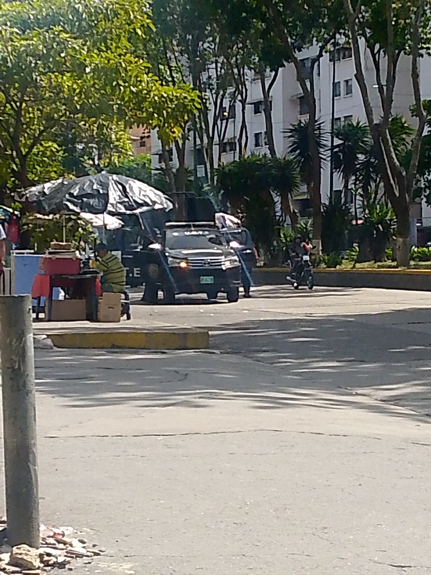 A street scene in Caracas, Venezuela, shows a black vehicle parked near a vendor's stand shaded by a large covering. A man wearing a striped shirt is seated near the stand. Another person stands near the vehicle. A motorcyclist rides on the road. Trees and buildings are in the background. Debris is scattered on the street near a light pole in the foreground.