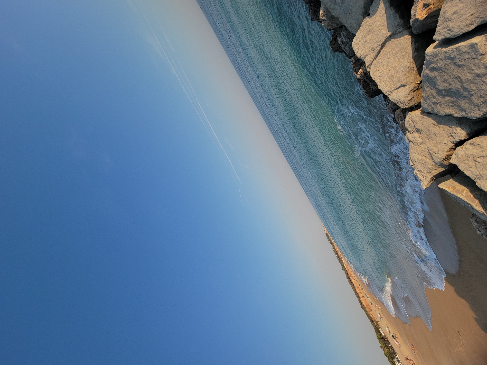 The image captures a wide-angle view of a beach and the ocean, likely in Quarteira, Portugal, given the distinctive coastline and architecture visible in the distance. The scene is dominated by a clear, bright blue sky taking up the upper portion of the frame. A faint, long white contrail from an aircraft stretches across the sky.
The ocean is a vibrant turquoise, with gentle waves lapping against the shore. In the foreground, large, rough-hewn rocks form a breakwater or natural barrier. These rocks are weathered and vary in size, with some casting distinct shadows on others, indicating the sun is at an angle.
The sandy beach stretches along the left side of the image, appearing populated with several small figures of people scattered across its width. Some individuals are lying on the sand, while others appear to be walking or standing. In the background, along the edge of the beach, a line of low-rise buildings and trees are visible, characteristic of a coastal resort town. The overall impression is one of a tranquil summer day at the seaside.