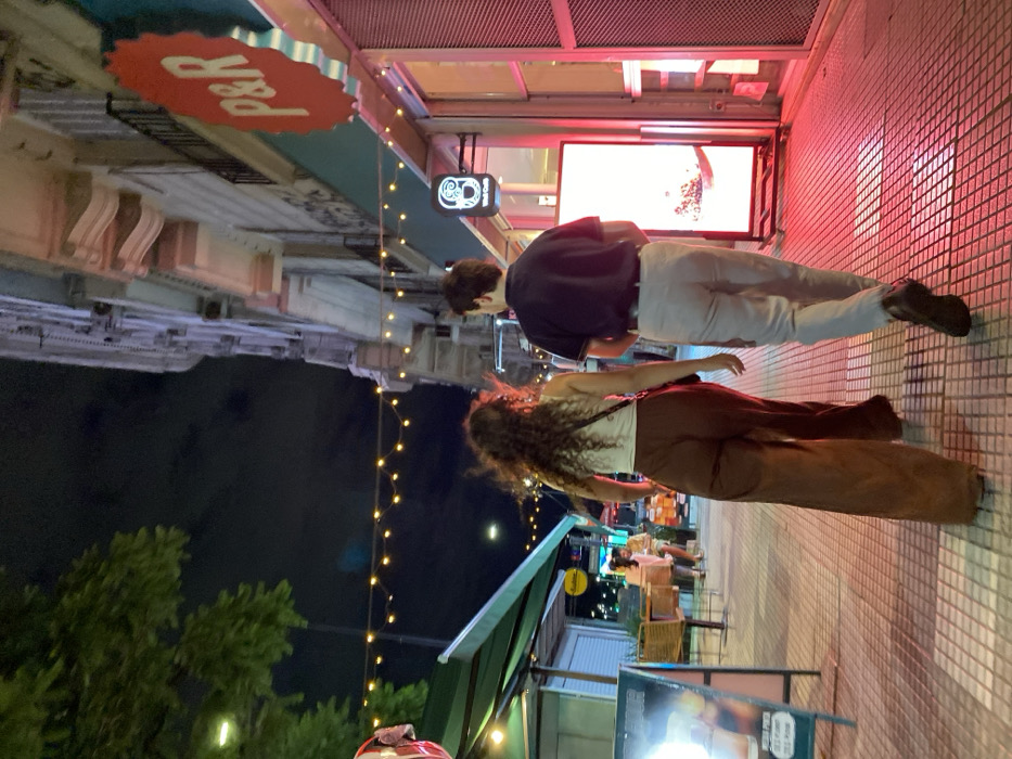A street scene at night depicts a couple standing on a tiled sidewalk, illuminated by a red glow. The woman has curly hair, a light-colored top, and brown pants. The man wears a dark shirt and light-colored pants. Overhead, buildings feature signs and glowing screens. String lights add to the ambiance. Trees and outdoor seating are visible in the background. The perspective is rotated, giving the impression that the scene is viewed from the side.
