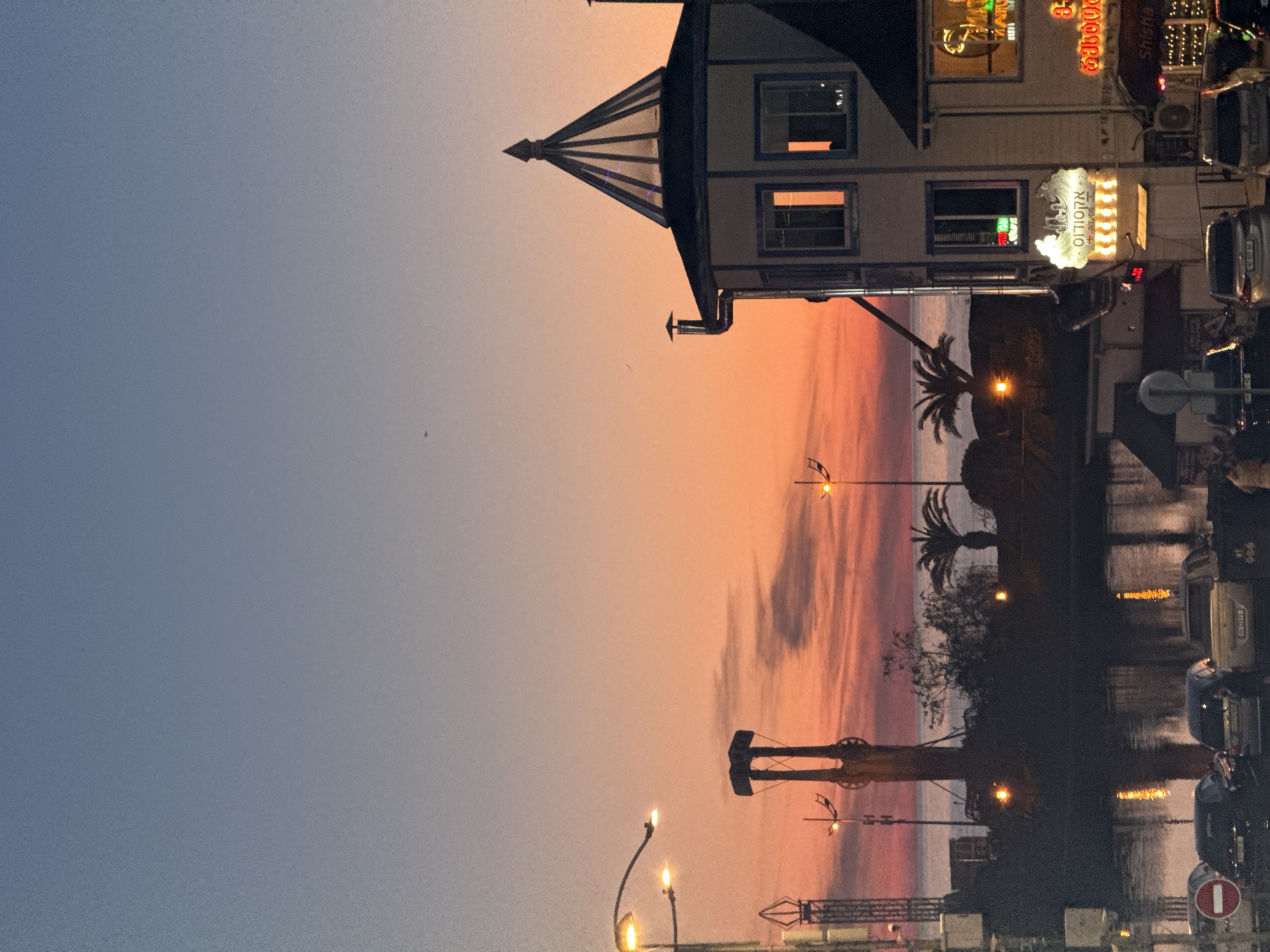The image depicts a sunset in Batumi, Georgia, likely near the seafront. The sky transitions from blue to orange, with reflections visible on the water. Silhouetted palm trees and a portion of a pier are visible against the sunset. A building with a peaked roof and lit signs, including Hebrew text and the word "Shisha," stands to the side. Parked cars and streetlights are also present, suggesting a bustling area.