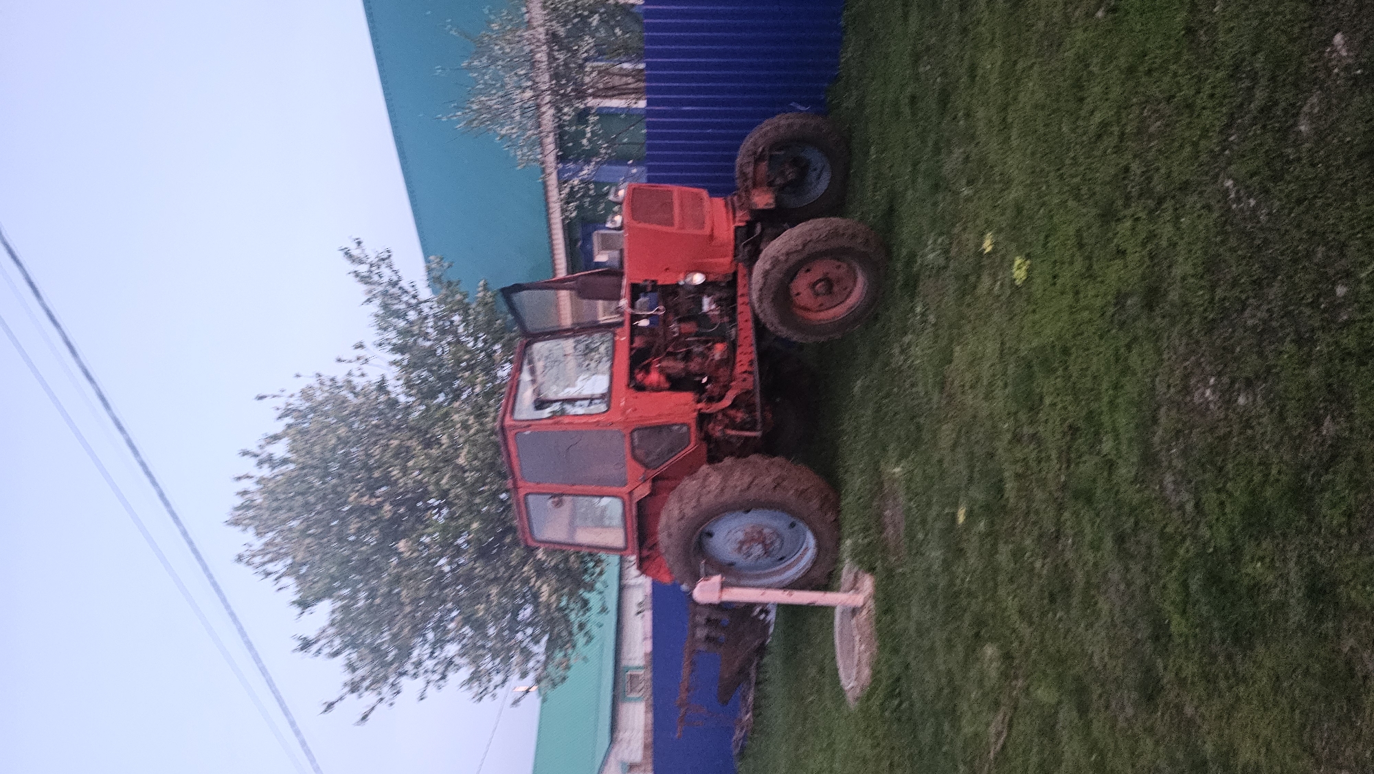 The image shows a red tractor parked on a grassy area, likely a lawn or field. The tractor has an open cab, revealing the engine and interior. It appears to be an older model. Behind the tractor is a blue corrugated fence, and a tree is situated near the cab. The setting appears to be outdoors. The location is described as Yangiskain, Russia.