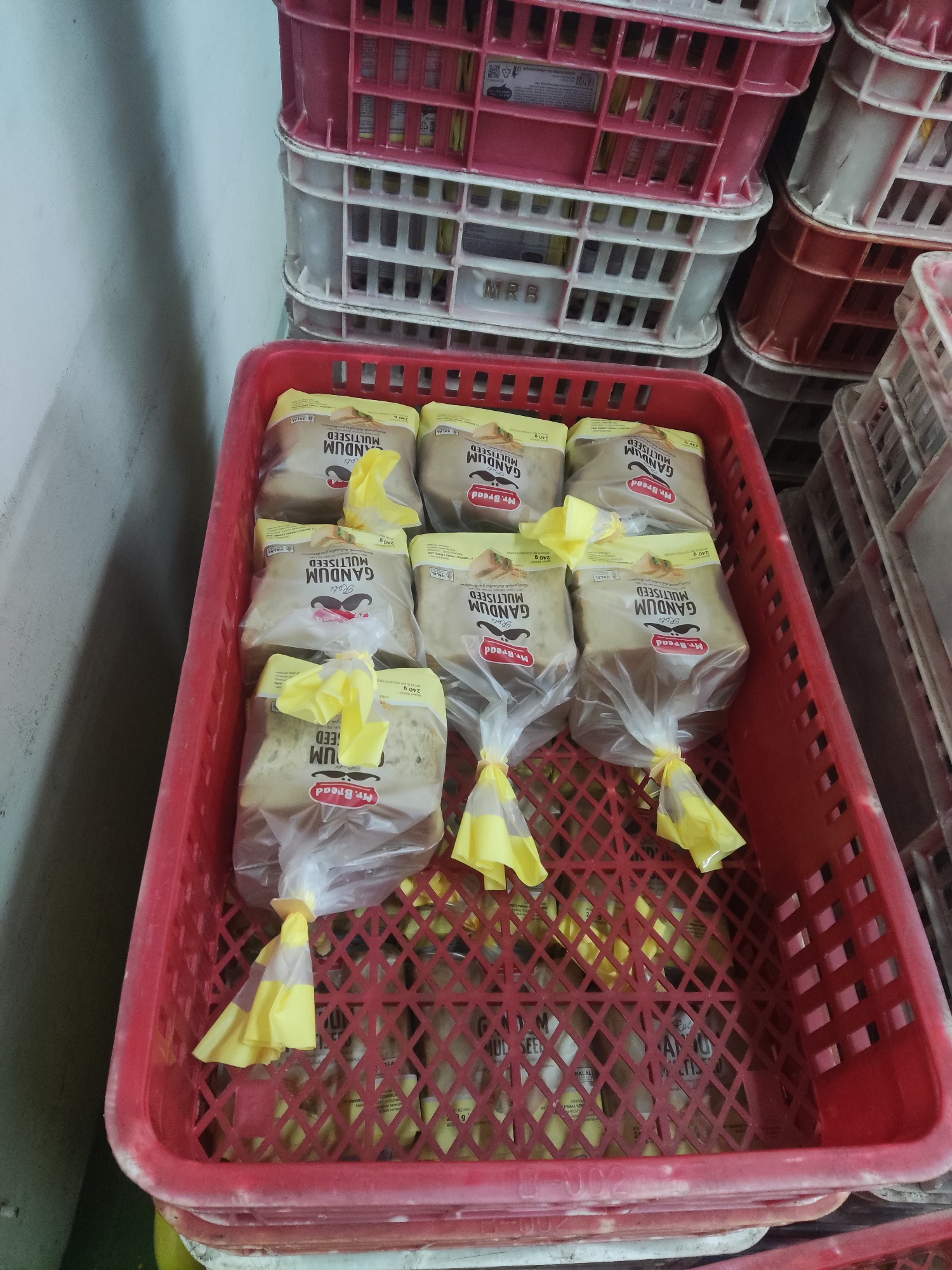 A red plastic crate contains several individually wrapped loaves of bread. Each loaf is encased in clear plastic packaging tied at the bottom with a yellow tie, with a yellow ribbon or bow above it. The bread packaging features text including "GANDUM MULTISEED", "Roti", and "Mr. Bread". The crate is situated on top of other crates, one of which is white and has the marking "MRB". The background shows a light blue wall and stacks of additional red and white crates. This scene is located in Trihanggo, Indonesia.