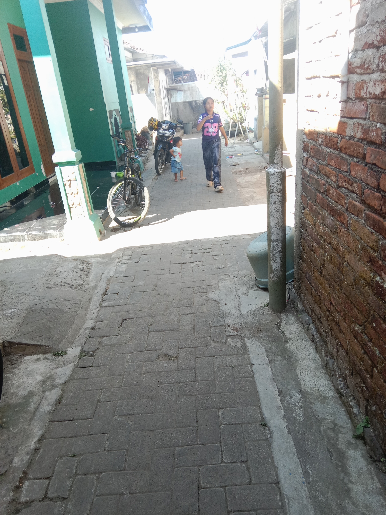 In the city of Dangkel, Indonesia, a paved alleyway is illuminated by bright sunlight. On the left, a turquoise and white building with wooden doors stands adjacent to the alley. In the middle ground, a young woman in striped pants and a purple shirt with a graphic design is walking away from the camera, while a small child in a blue outfit walks ahead of her. A bicycle with a white-rimmed wheel is parked on the left side of the alley, near a black motorcycle. The right side of the alley is bordered by a brick wall, with a concrete pole and a green trash bin visible in front of it. The ground is paved with rectangular grey bricks, with some patches of concrete and vegetation growing between the stones.