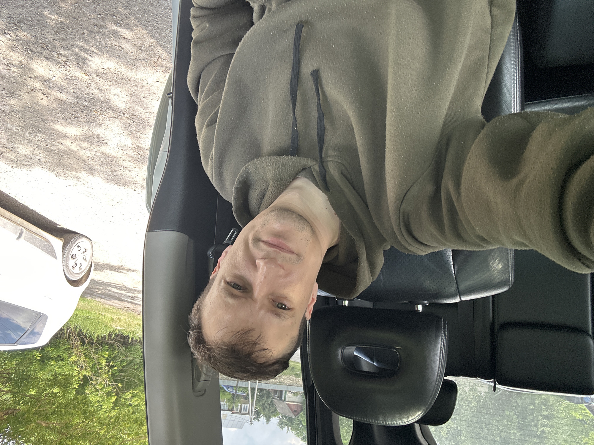 In Makiv, Ukraine, a man in his late twenties or early thirties, with light skin and short brown hair, is taking a selfie inside a car. He is wearing a dark olive green hooded sweatshirt. The car's interior is visible, with black leather seats and headrests. Through the car window, a rural landscape with houses, trees, and a white car parked on a dirt road can be seen. The lighting suggests it is daytime.
