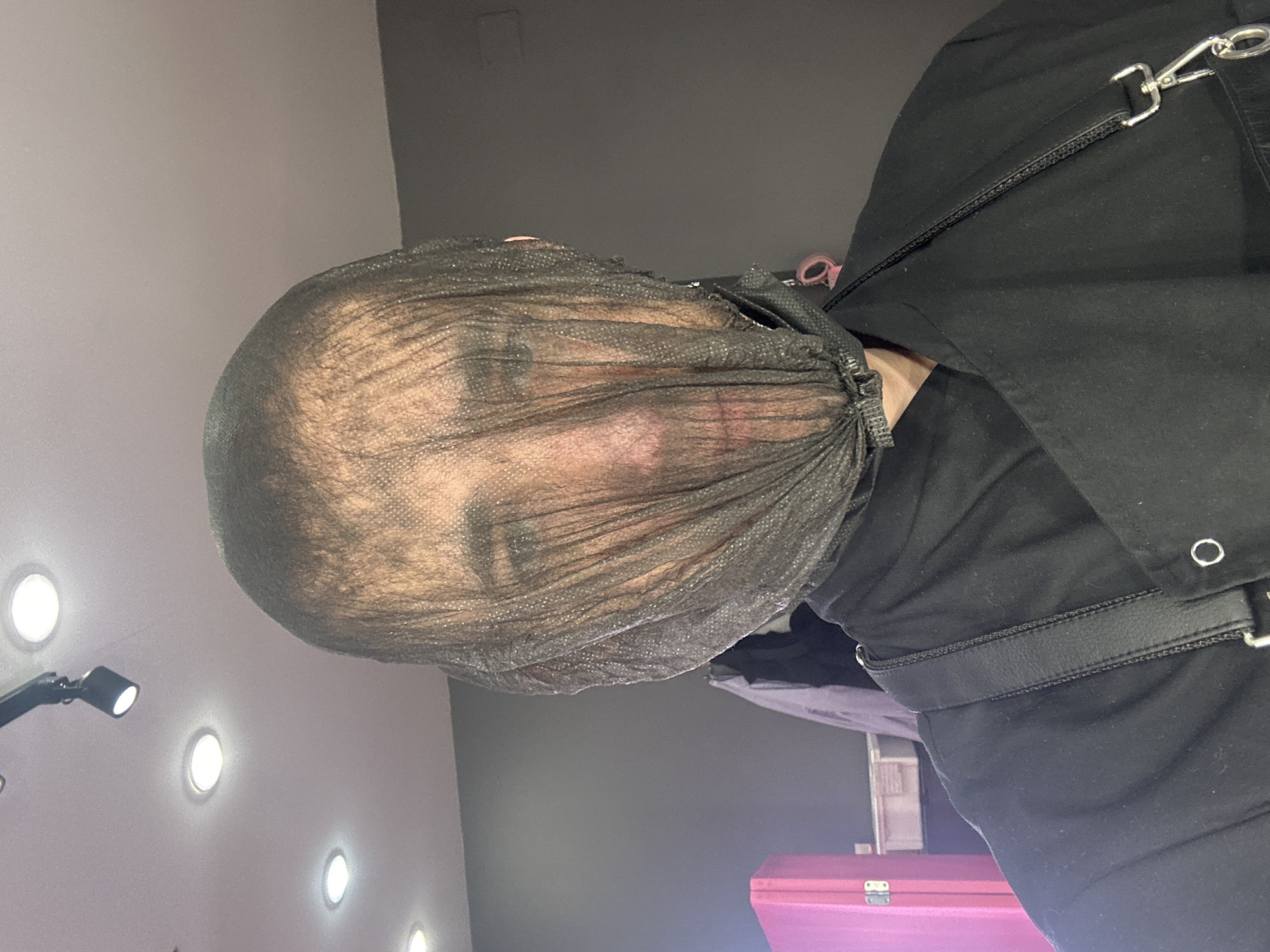 A person's head is enclosed in a black net. The person is wearing black clothing with a leather belt and metal hardware. The background appears to be a room with multiple round lights. A pink object is visible in the lower part of the image. There is no indication of the city of Saratov, Russia.