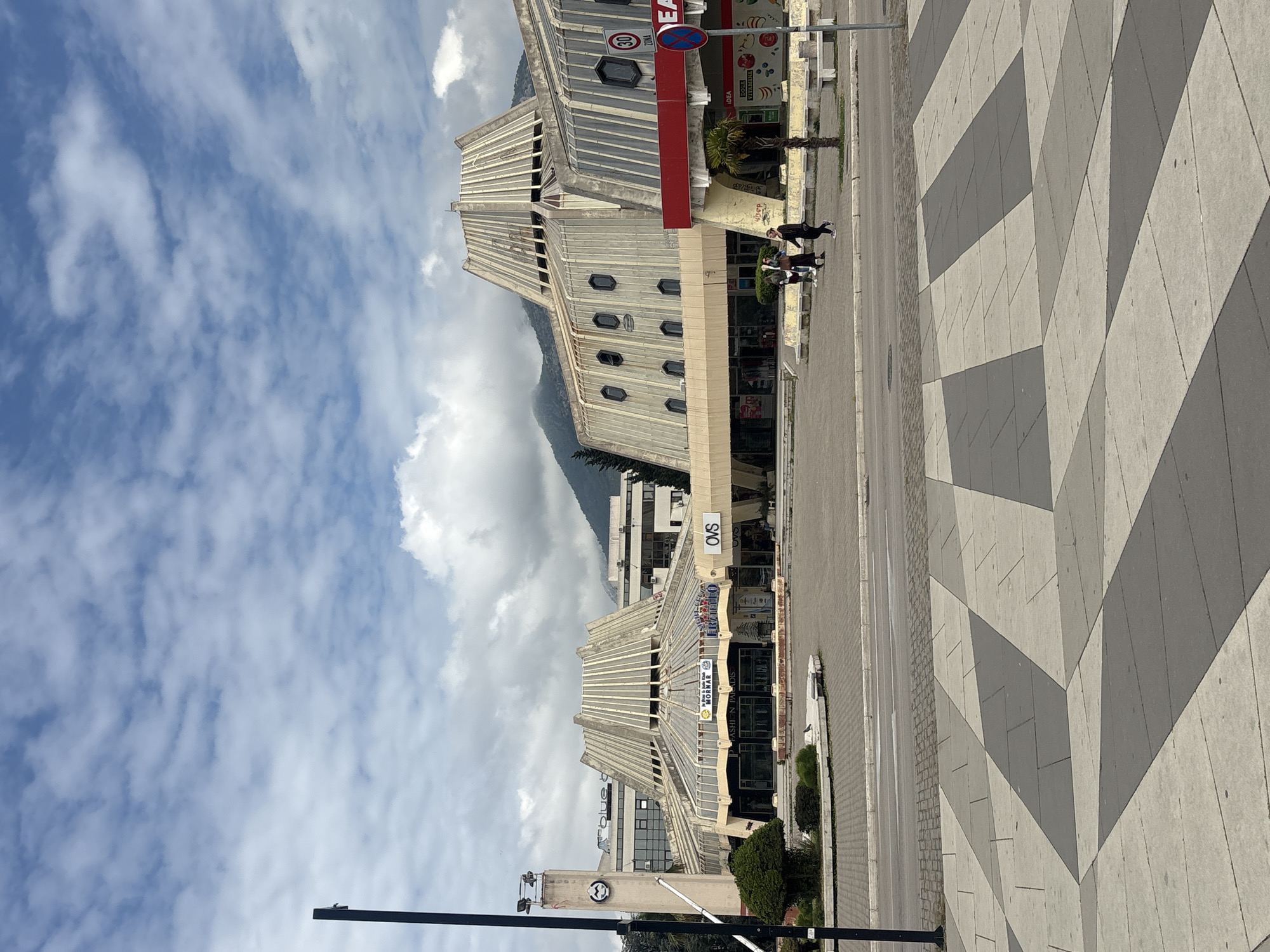 The image depicts a daytime scene in Bar, Montenegro, focusing on the architectural design of a building. The building has a distinct brutalist style with repetitive, angular concrete elements, rising several stories. There are shops along the street level. The sky is partially cloudy. The sidewalk is paved in a geometric, zigzagging pattern. There are few pedestrians visible on the sidewalk. A mountain is visible in the background behind the building. There are street signs and utility poles are present.