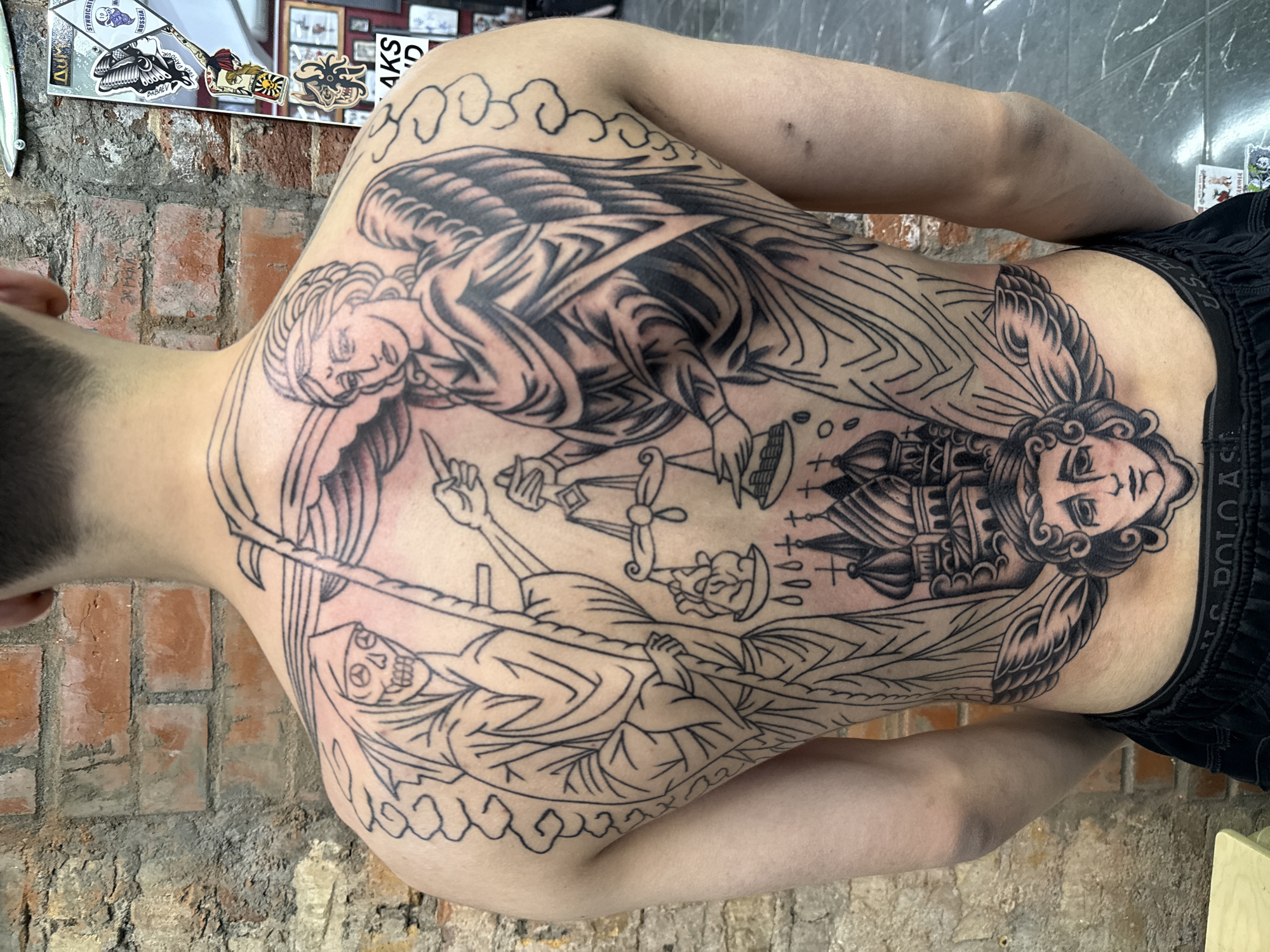 The image depicts a tattoo on a person's back. The tattoo features two winged figures, one at the top and one at the bottom, with an image of the Grim Reaper in the center. The top figure is holding a scale. Between the two winged figures is a tower or building. The person wearing the tattoo is standing in front of a brick wall. The location in the city of Moscow, Russia is not apparent from the image.