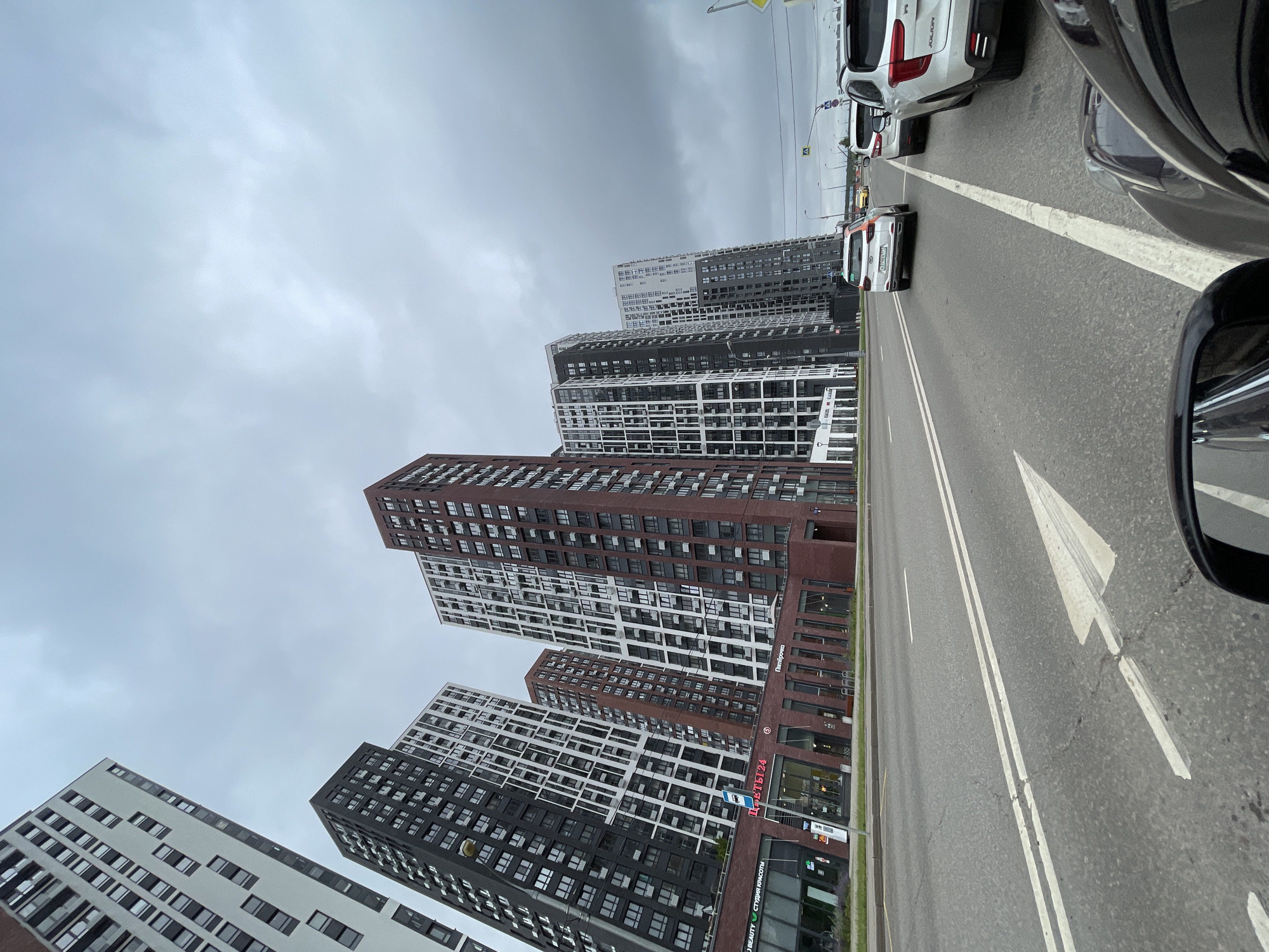 The image depicts a street scene in Moscow, Russia, characterized by modern, high-rise apartment buildings lining the left side of a multi-lane road. The architecture of the buildings is varied, with some featuring dark facades with white geometric patterns and others displaying reddish-brown brickwork. The sky is overcast and appears to be filled with dense, grey clouds, suggesting a cloudy or possibly rainy day.
In the foreground, the asphalt road is marked with white lane lines and directional arrows. Several cars are visible on the road, indicating traffic. On the right side of the road, a line of vehicles is stopped or moving slowly, suggesting potential congestion. One white SUV with a black roof is prominently featured in the foreground on the right.
The buildings on the left have commercial spaces at their ground level, with visible storefronts and signage. One sign appears to read "ЦВЕТЫ24" (FLOWERS24) and another "Пятерочка" (Pyaterochka), which is a common Russian supermarket chain. The overall impression is of a contemporary urban environment.